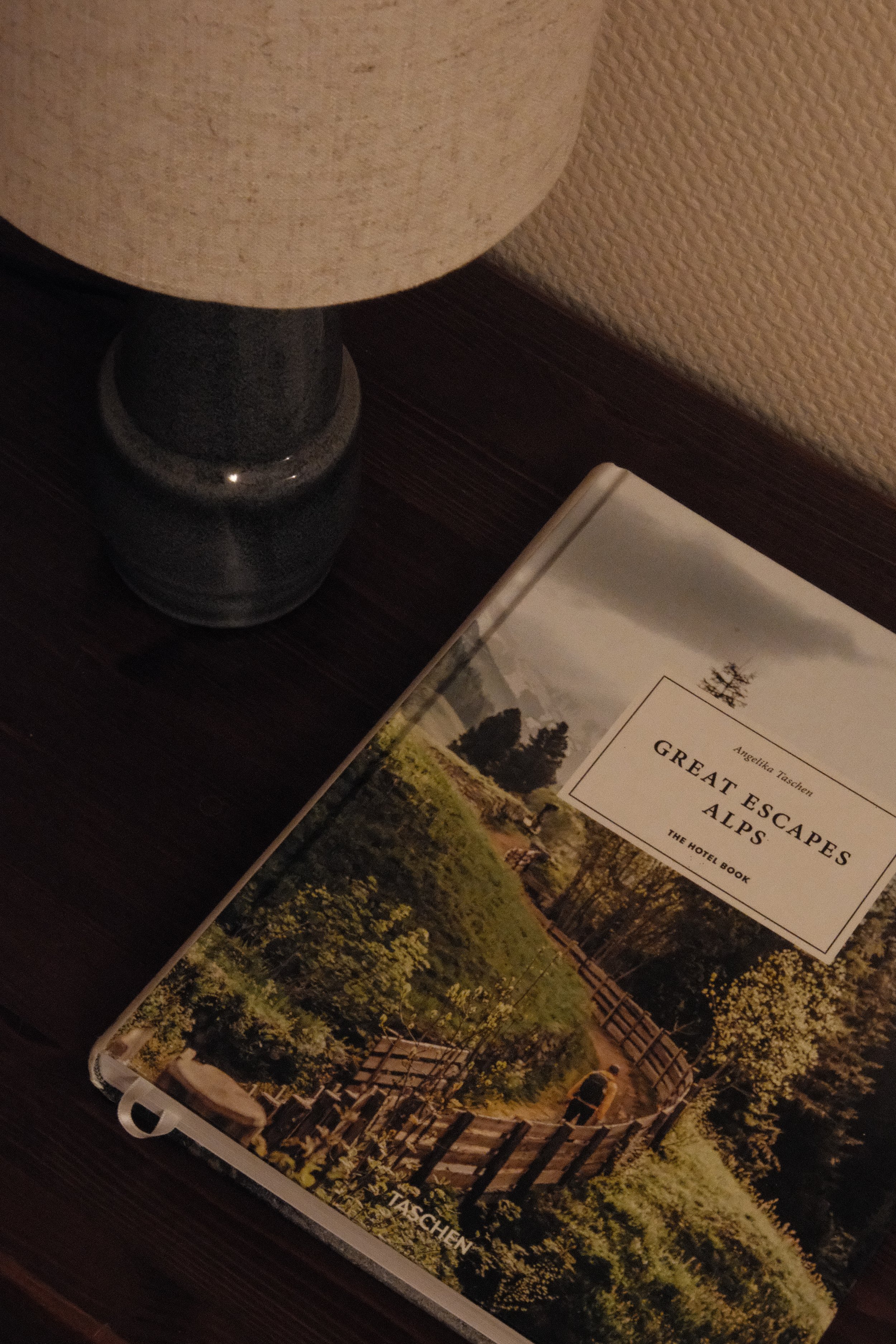Reiseführer mit dem Titel 'Great Escapes Alps', liegend auf einem dunklen Holztisch neben einer Tischlampe.