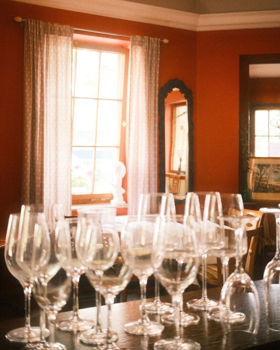 Mehrere leere Weingläser auf einem Tisch in einem rustikalen, gut beleuchteten Raum mit braunen Wänden, hohen Fenstern und Vorhängen.