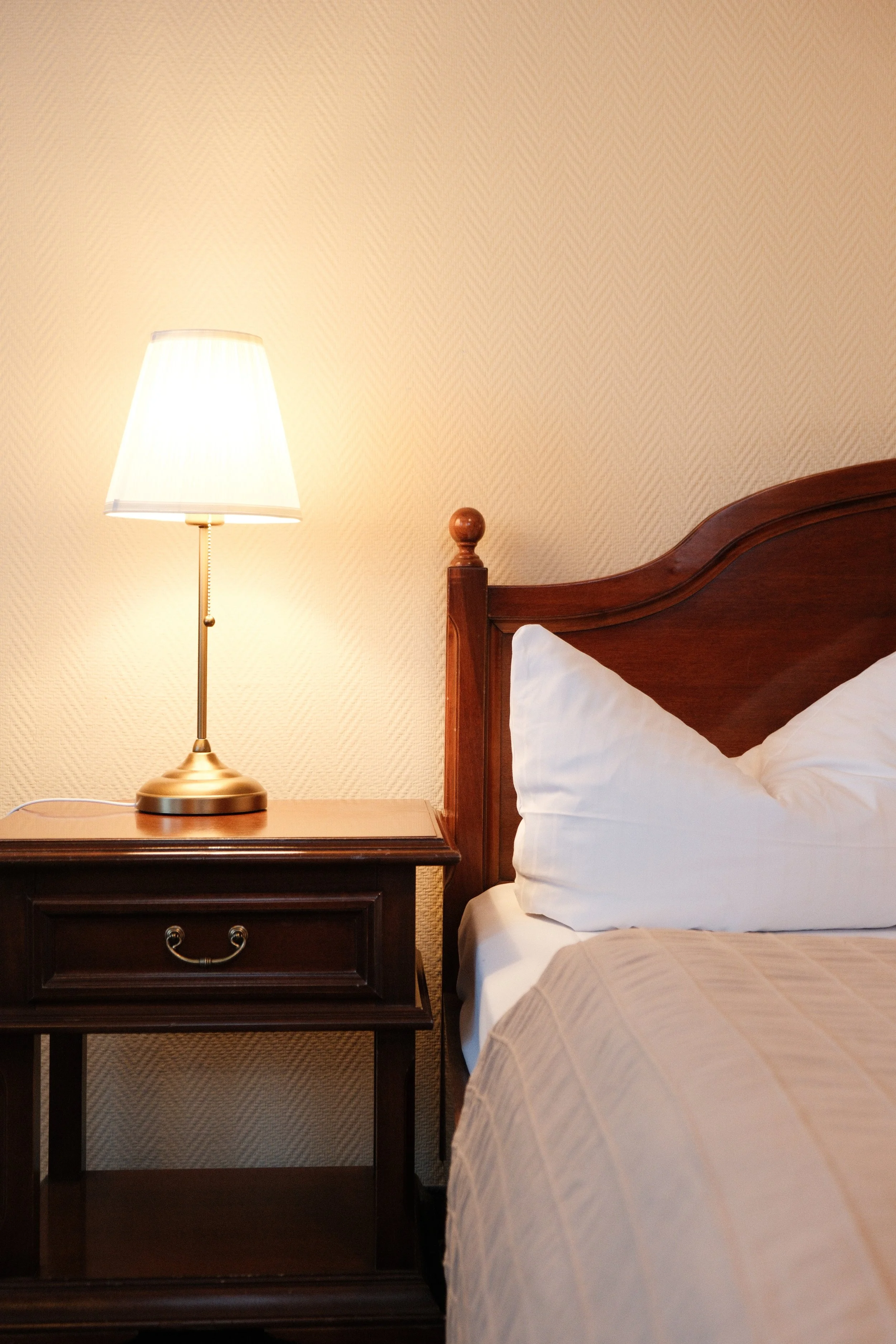 Ein Nachttisch mit einer Lampe steht neben einem Holzkopfkissen auf einem Bett, in einem Schlafzimmer mit cremefarbener Wand.