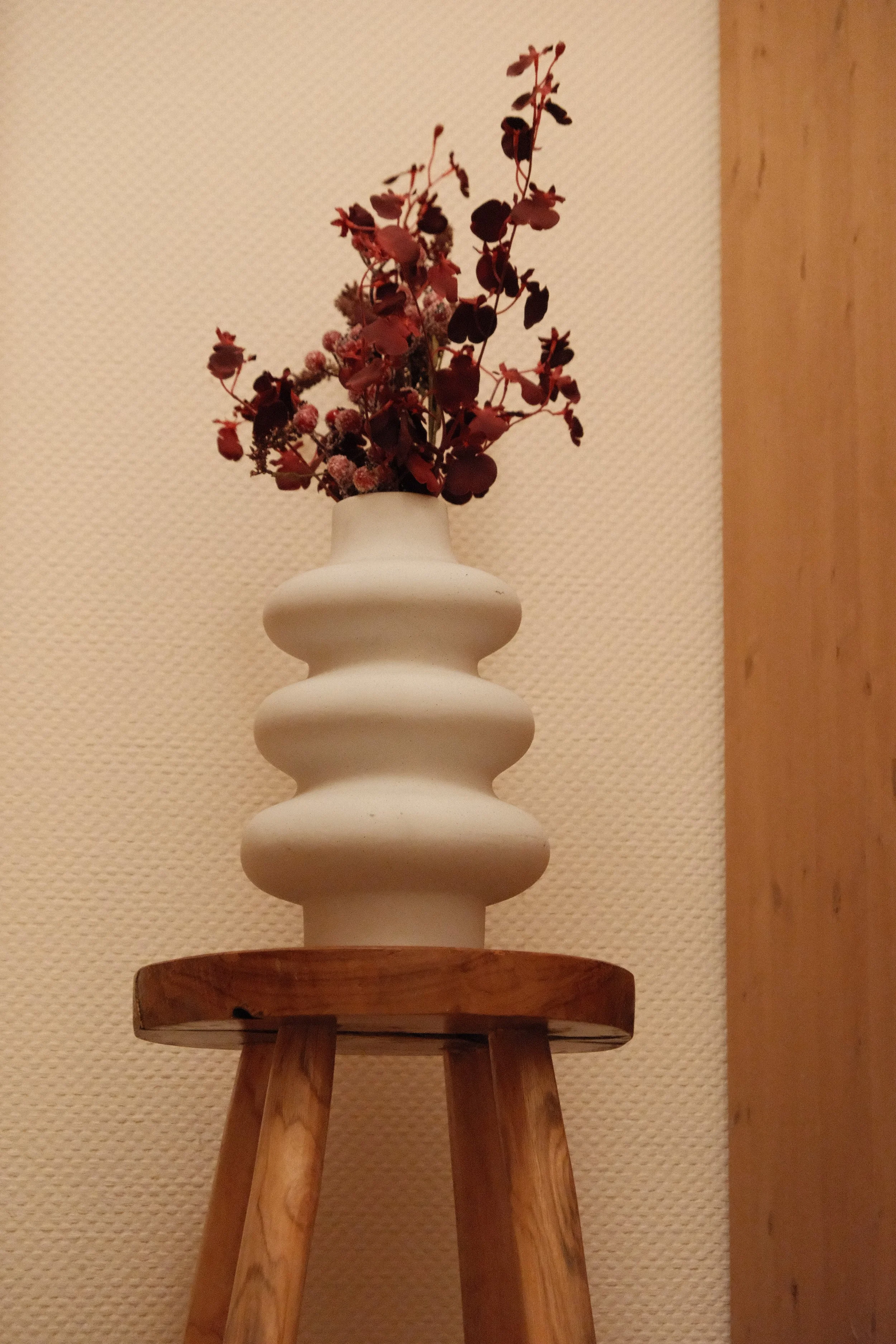 Vase mit getrockneten roten und pinkfarbenen Blumen auf einem Holzstuhl vor einer beige gemusterten Wand.