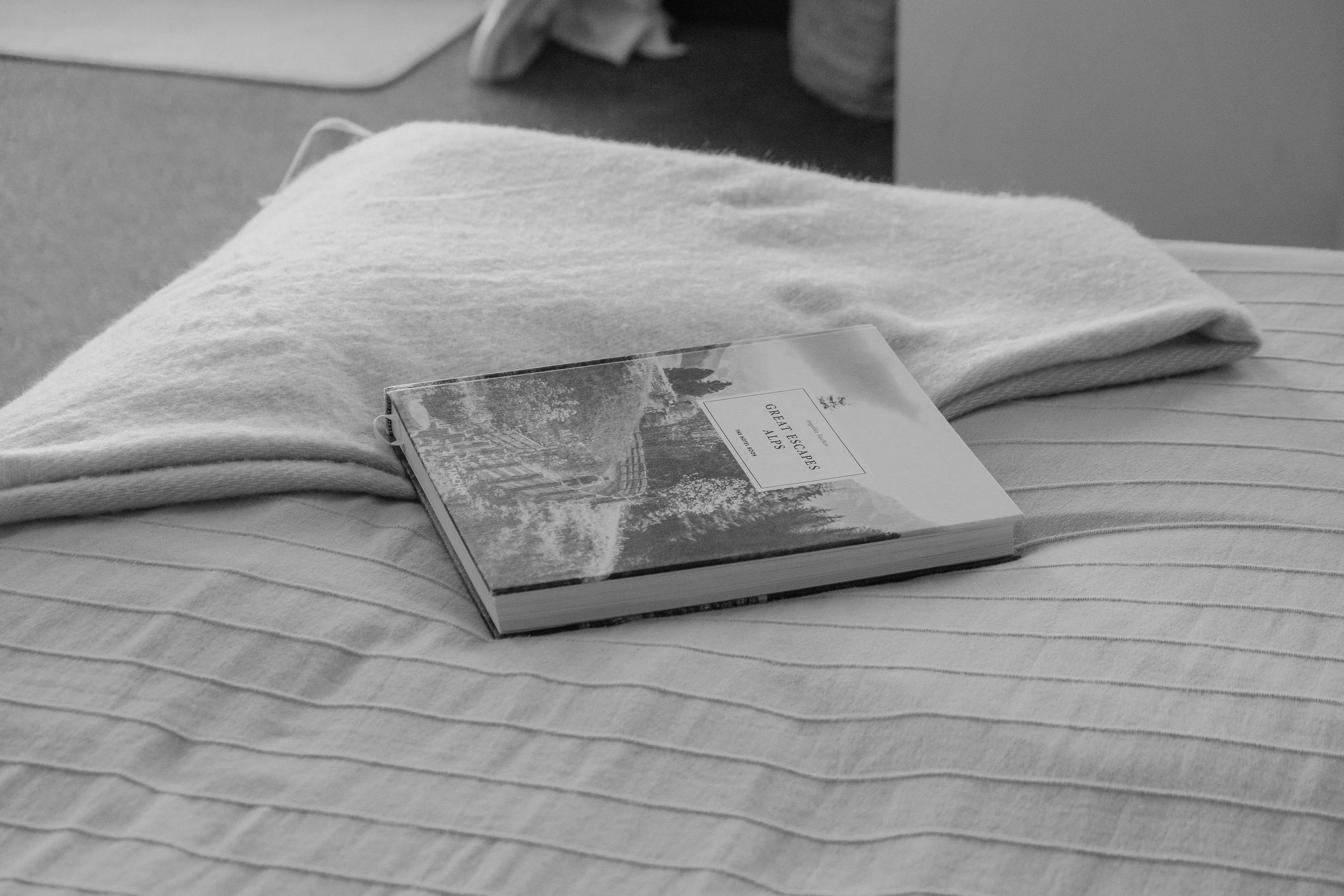 Schwarz-weiß-Fotografie eines Buchs namens 'Great Escapes Alps' liegend auf einem Bett, mit einer weißen, gestrickten Decke und einem Kissen im Hintergrund.