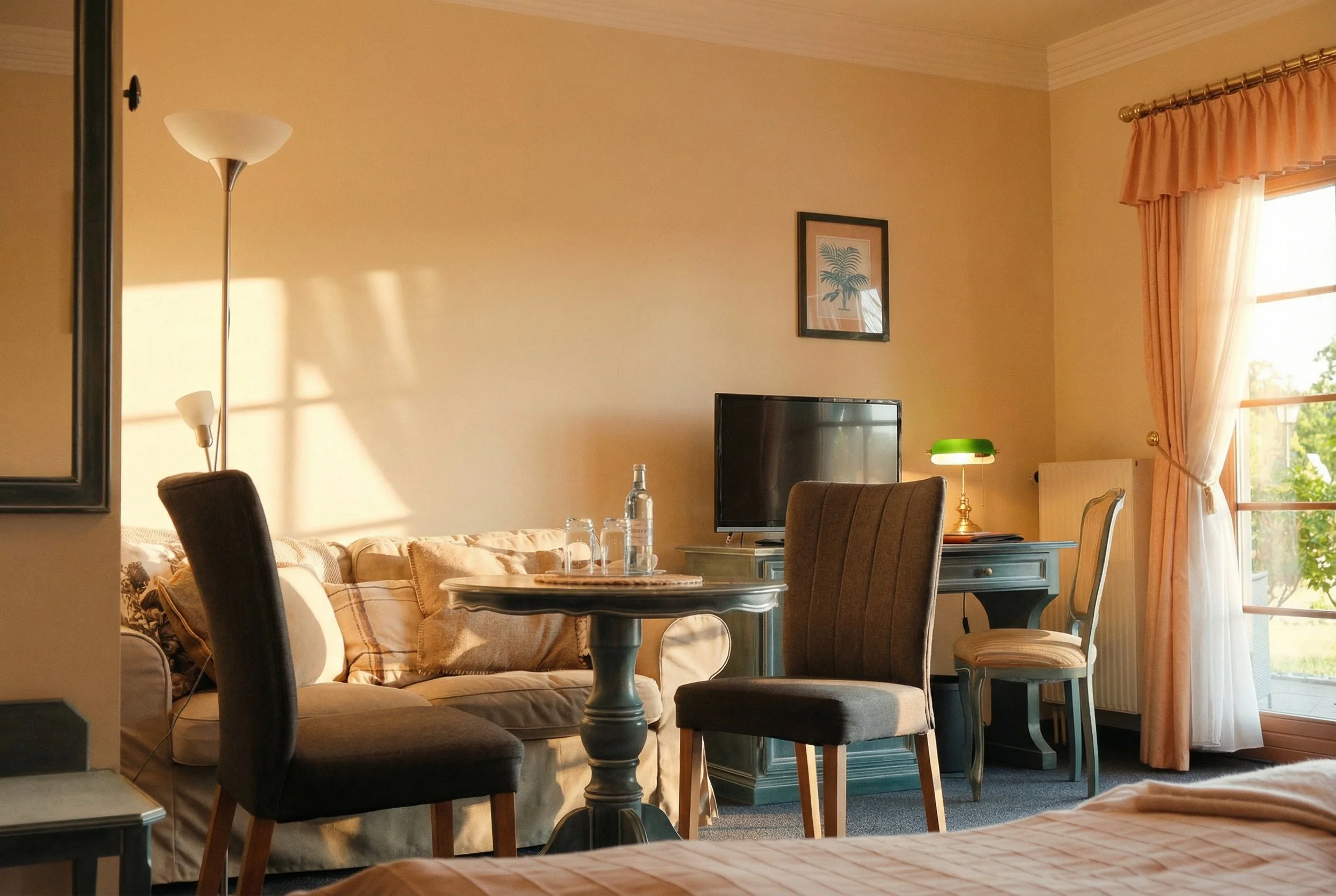 Ein Hotelzimmer mit Sofa, Stühlen, einem Fernseher, einem Tisch mit Wasserflaschen, und einem Schreibtisch mit einem grünen Lampen. Das Zimmer ist hell erleuchtet durch Sonnenlicht, das durch die Fenster fällt.