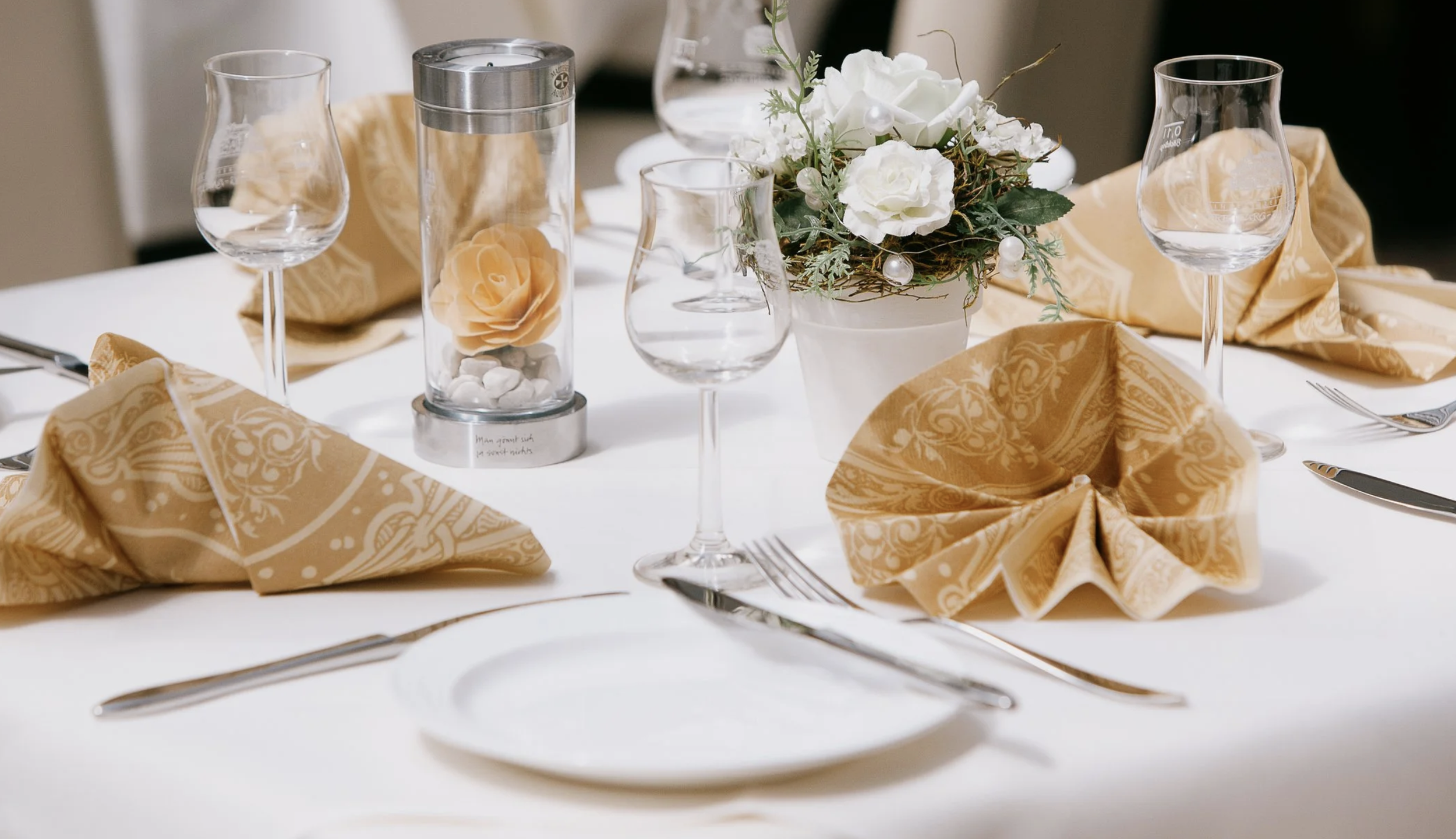 Elegant gedeckter Esstisch mit weißen Teller, goldenen Stoffservietten, Weingläsern, Kerze und Blumenstrauß in Weiß, auf weißem Tisch.