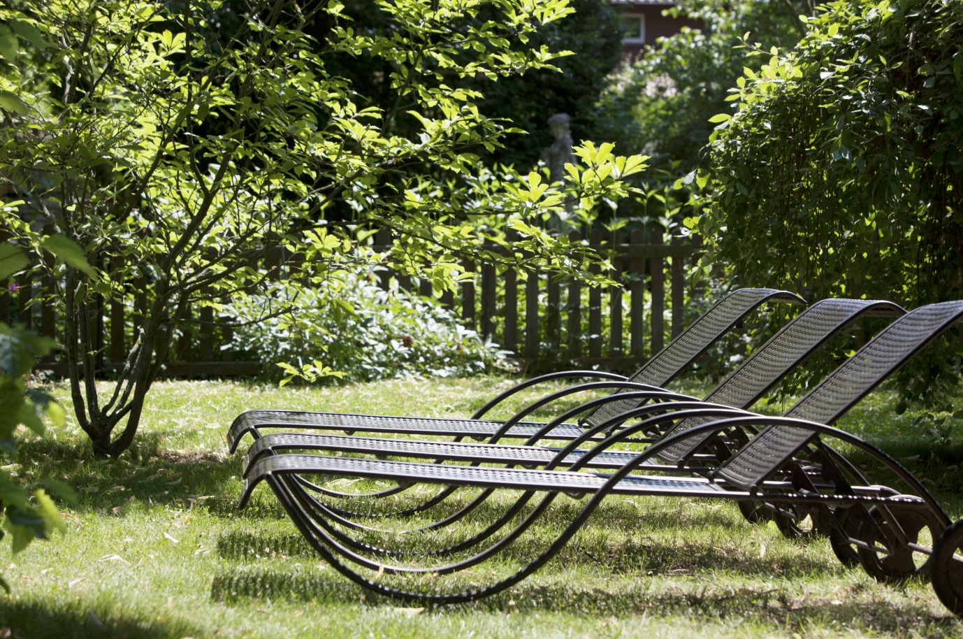 Drei mehrere Sonnenliegen aus Metall auf einer grünen Rasenfläche, umgeben von Büschen und Bäumen in einem Garten.