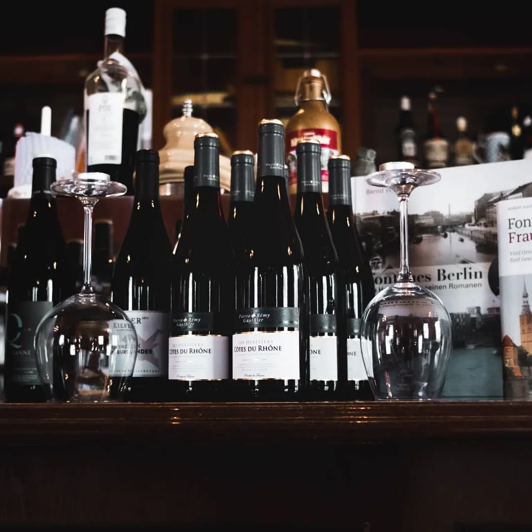 Mehrere Flaschen Rotwein auf einem Holzregal, umgeben von leeren Weingläsern. Es gibt auch Zeitschriften und eine Flasche mit schwarzem Etikett im Hintergrund.