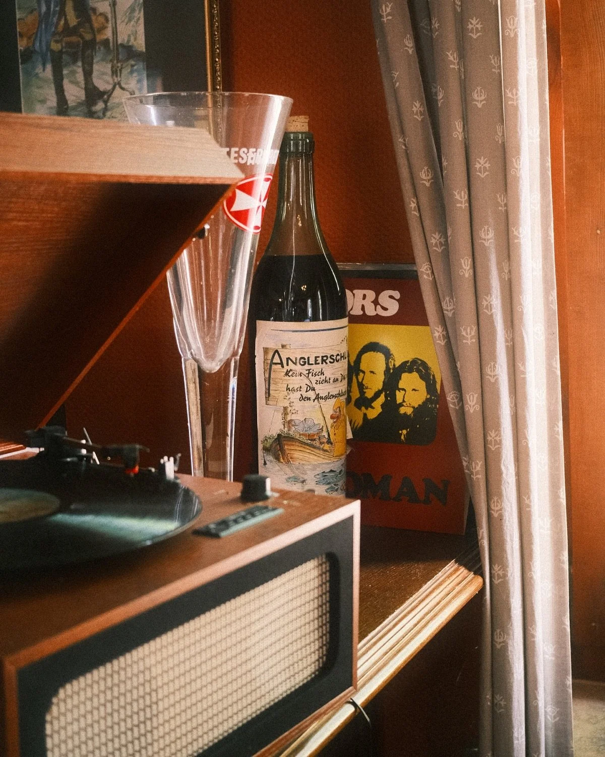 Ein stapelweise Aquarium-Flasche, ein Glas, ein Plattenspieler, ein Poster mit den Gesichtern von zwei Männern, ein Vorhang, und eine Schallbox auf einem Holzregal.