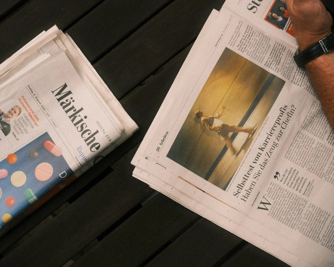 Zeitung 'Märkische' liegt auf einem dunklen Holztisch, daneben eine weitere Zeitung mit einem Foto eines Mannes, der auf einem Podium steht, mit einer Hand in der Hosentasche und einem Mikrofon in der anderen Hand.
