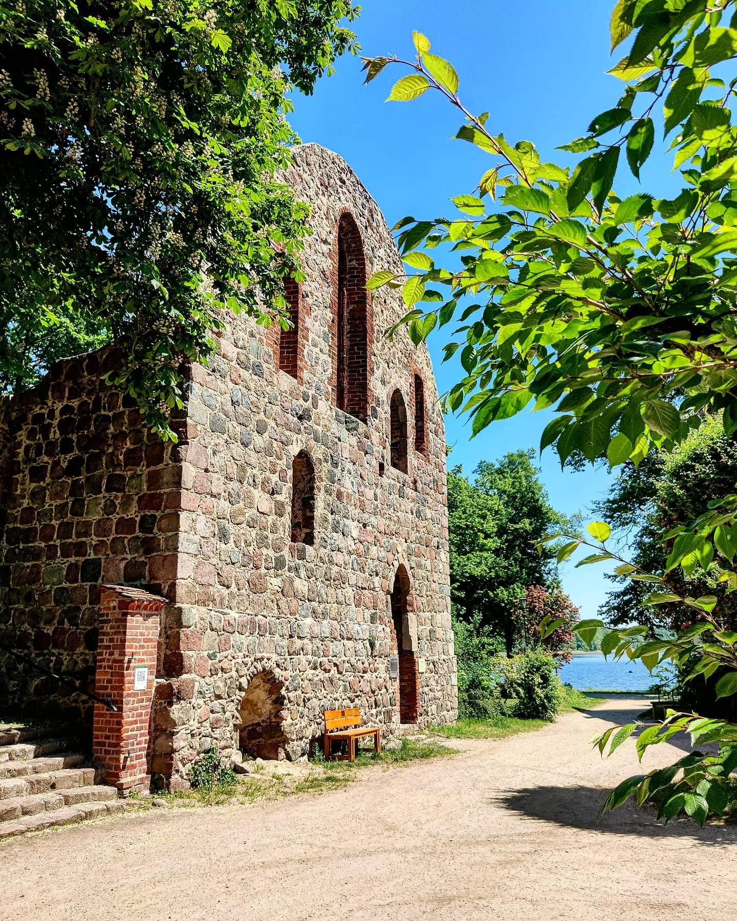 Alte steinerne Ruine an einem sonnigen Tag, mit Bäumen im Vordergrund und Wasser im Hintergrund.