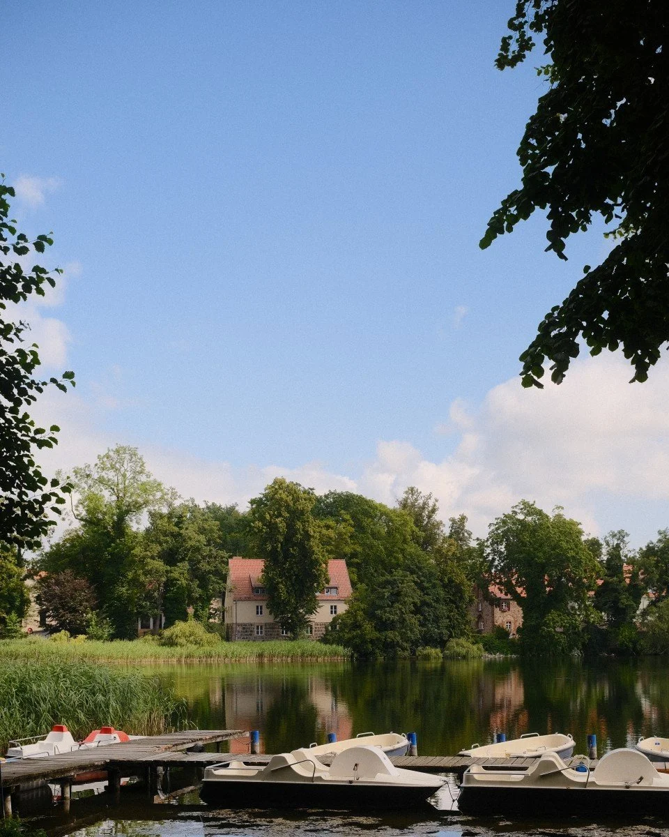 Ein ruhiger See mit mehreren weißen Tretbooten am Steg, umgeben von grünen Bäumen und Häusern am Ufer, bei sonnigem Wetter.