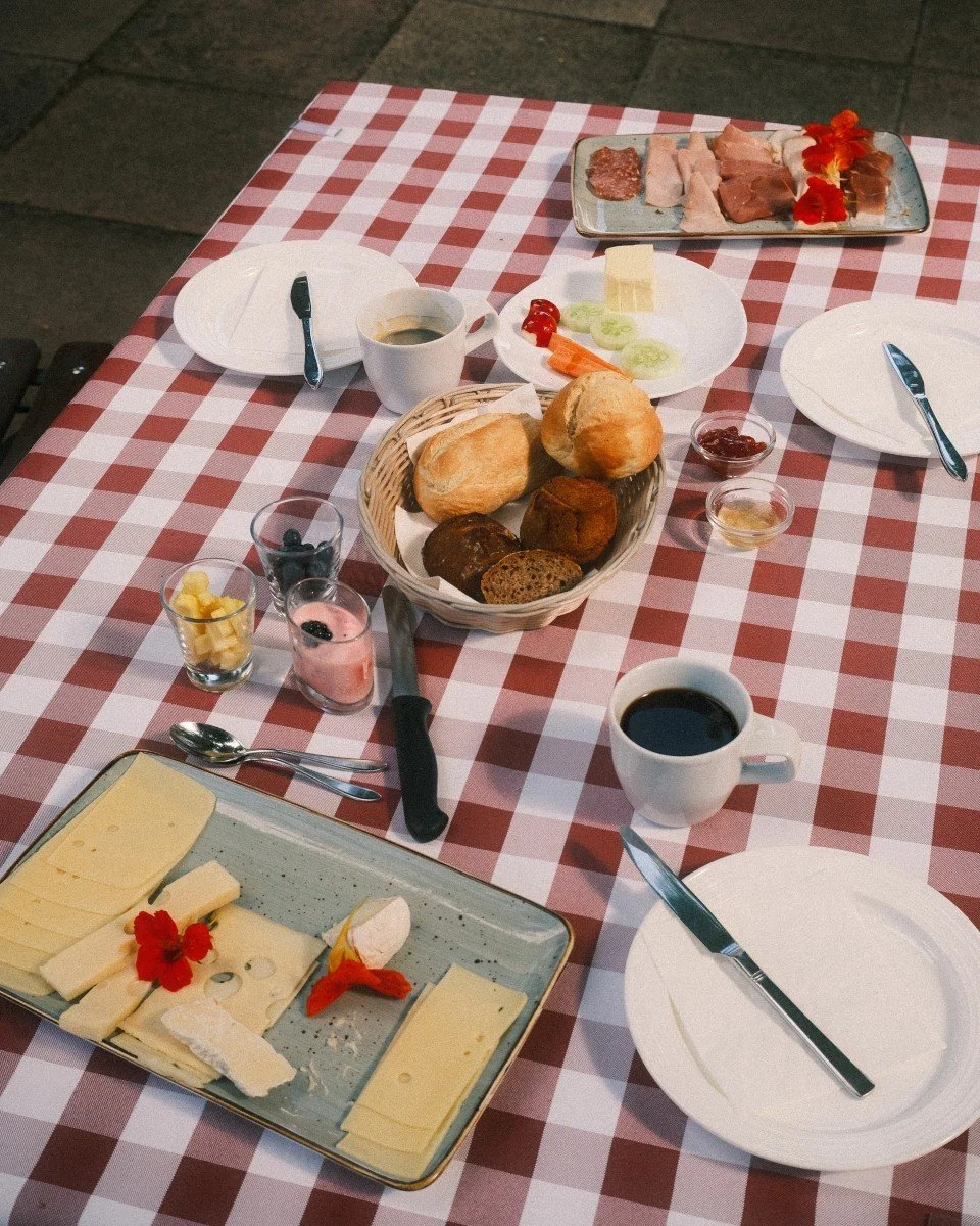 Frühstückstisch mit verschiedenen Broten, Käse, Aufschnitt, Marmelade, Kaffee und Joghurt auf einem rot-weiß karierten Tischtuch.