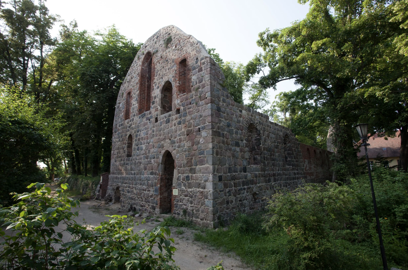 Fragment einer alten Steinmauer mit großen Fenstern, umgeben von Bäumen und Grünflächen im Freien.