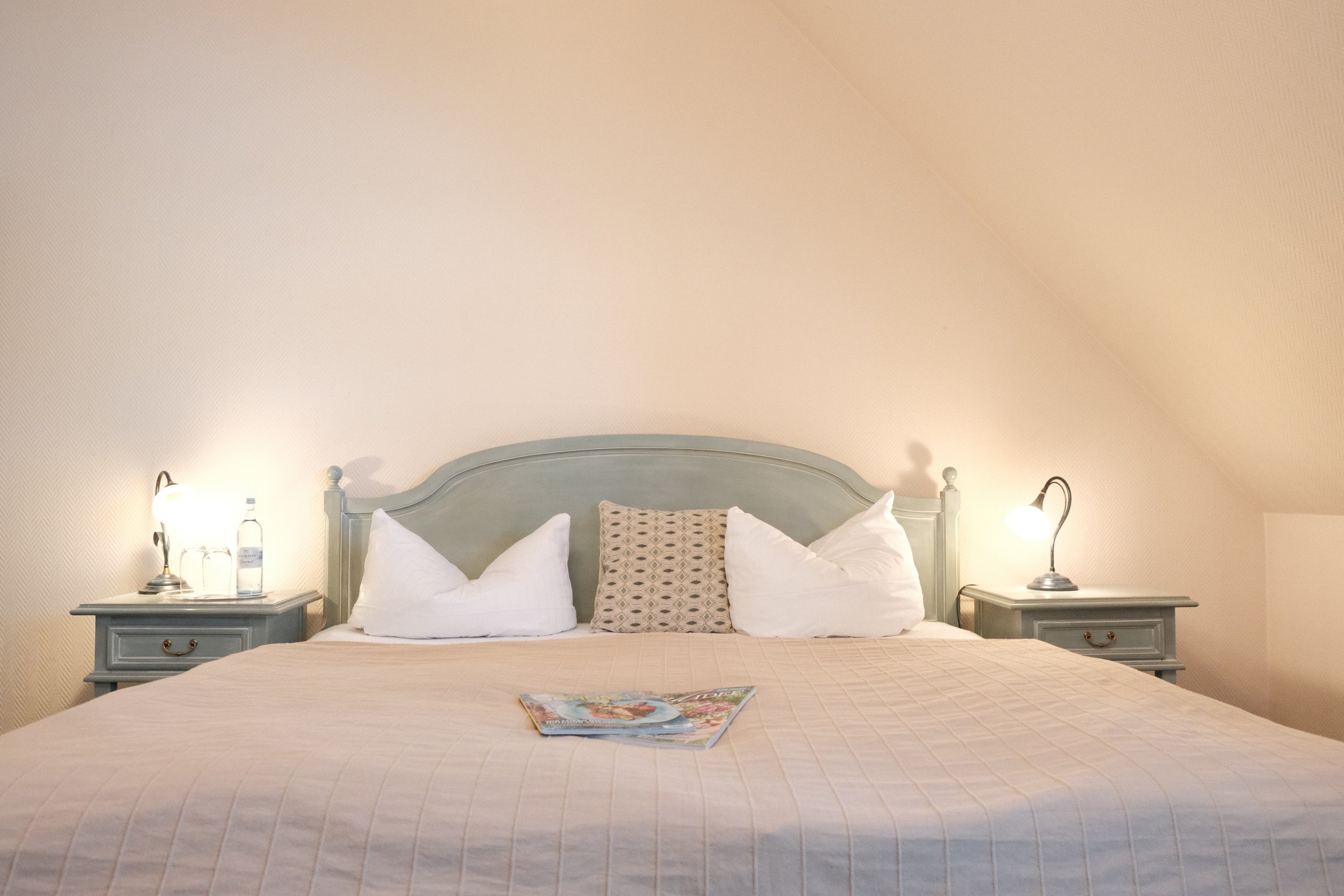 Bett mit weißen Kissen und Tagesdecken, Nachttische mit Lampen auf beiden Seiten, an einem Ende liegen Zeitschriften auf der Decke.