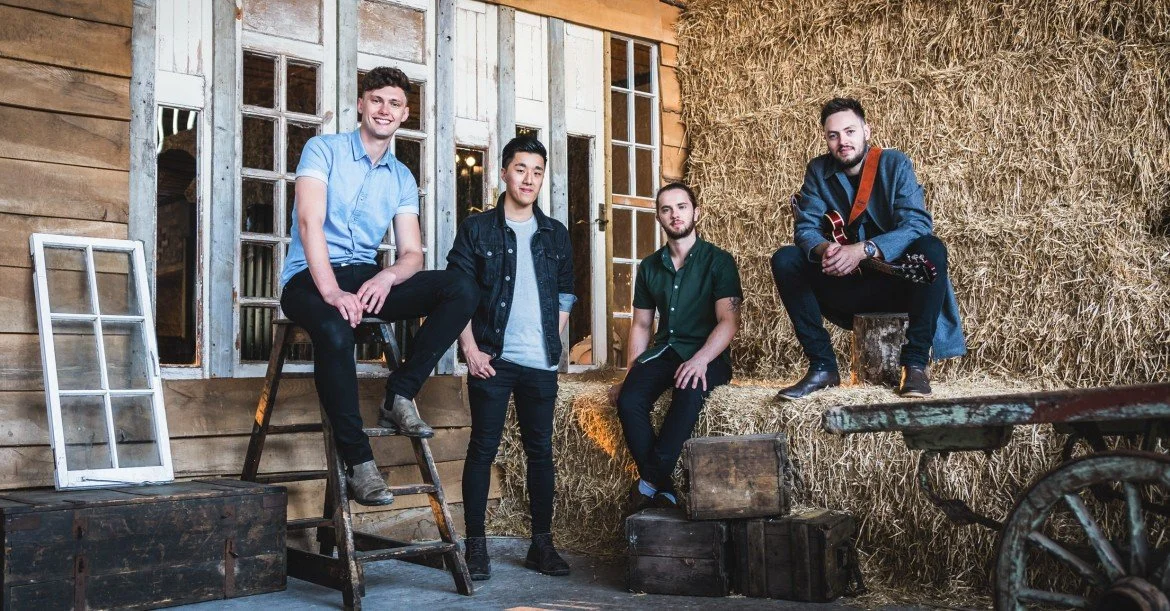 Four men in a rustic barn setting, with hay bales and wooden crates, some sitting and others standing, one holding a guitar.