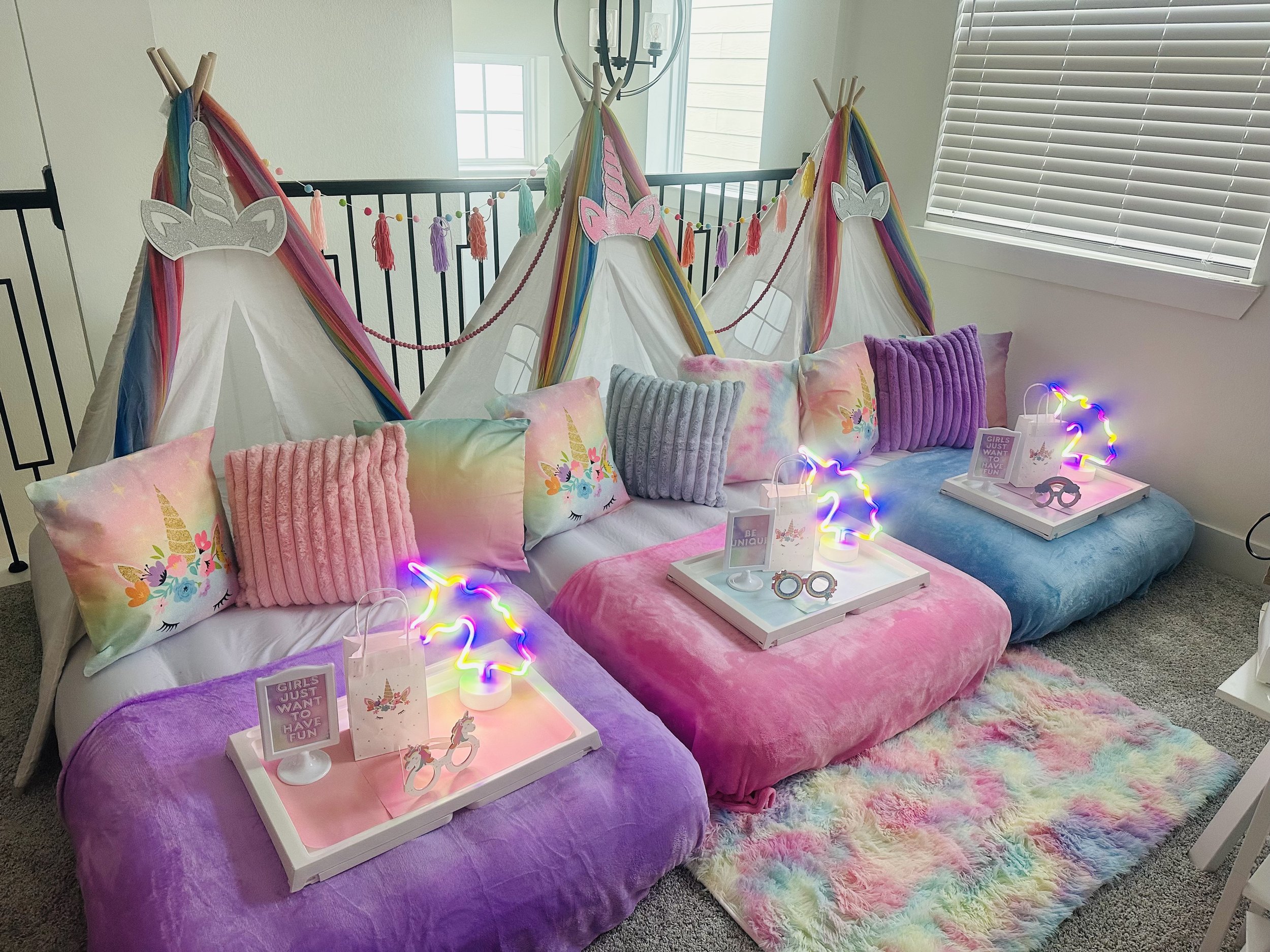 Colorful girls' sleepover setup with unicorn themed tents, rainbow and unicorn pillows, and pink, purple, and blue velvet bean bag chairs, each with a decorative tray, pink and purple unicorn string lights, and a rainbow fluffy rug.