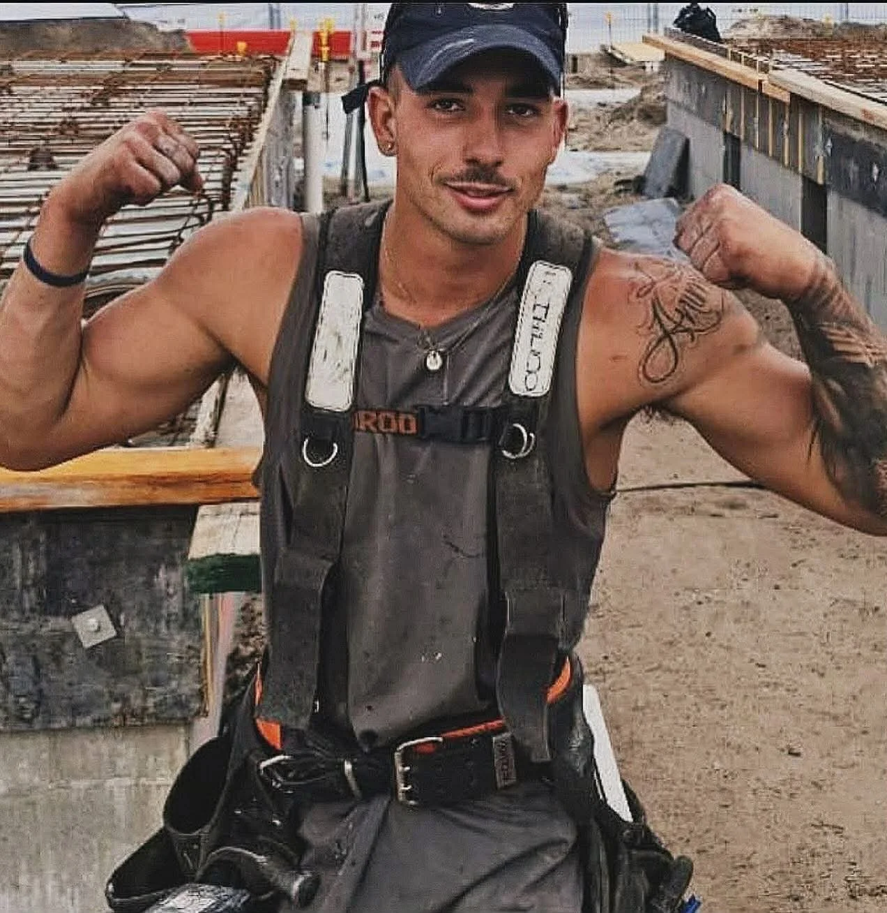 A young man wearing a work tool belt flexes his biceps at a construction site, smiling and showing tattoos on his arms.