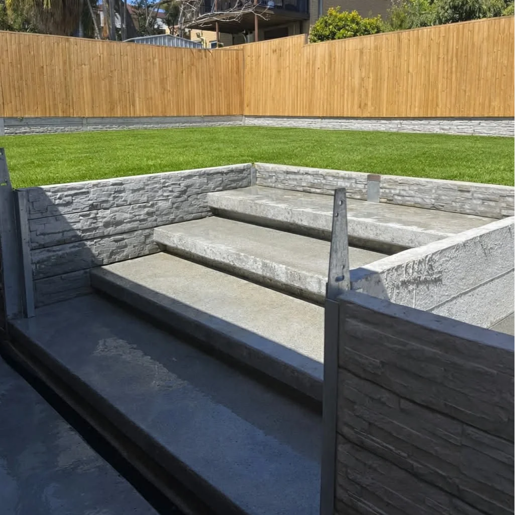 Concrete steps leading to an elevated yard with green grass, surrounded by a stone retaining wall and a wooden fence in the background.