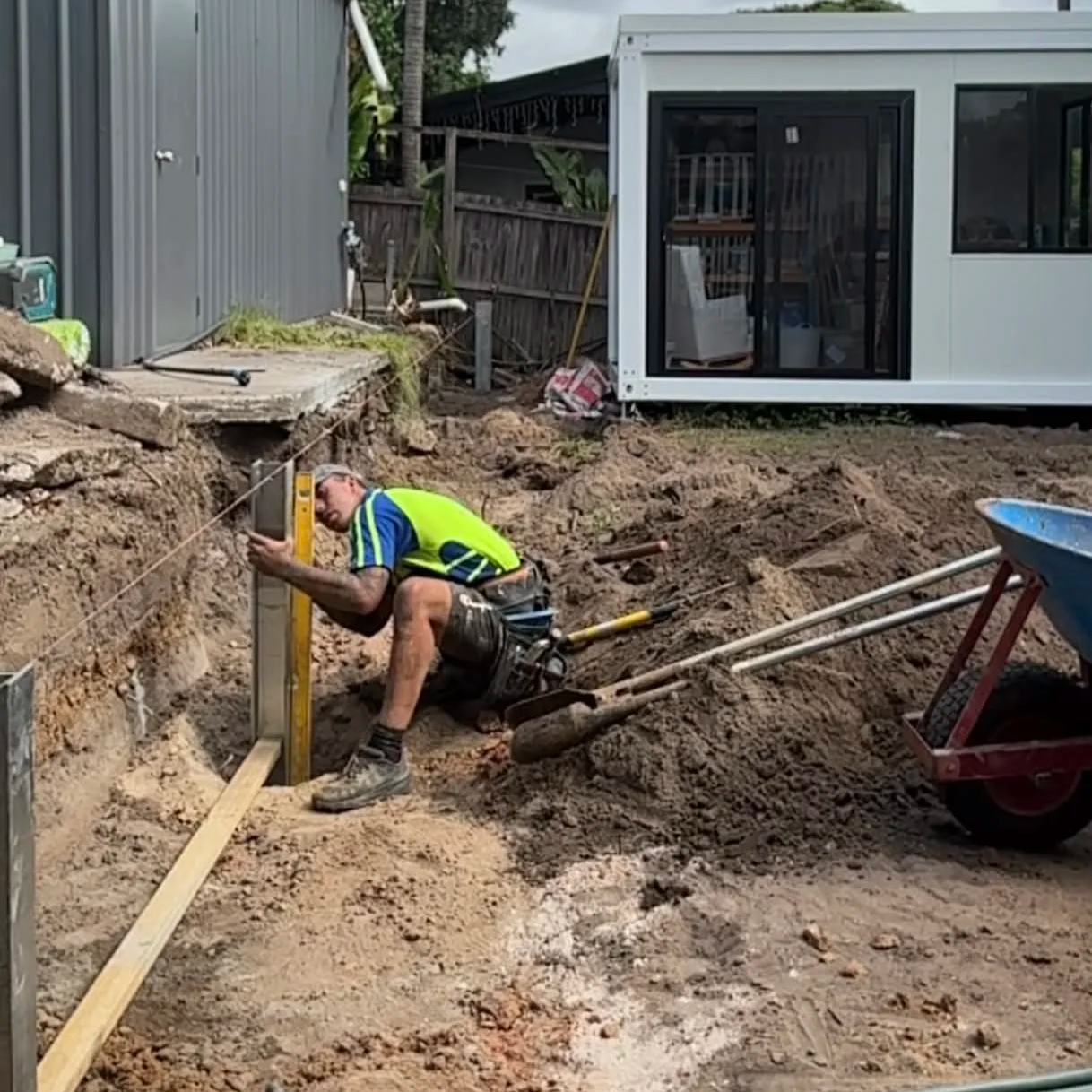 Construction worker kneeling in a dirt excavation, using a level tool on a vertical wooden post, with construction tools and equipment around, including shovels, a wheelbarrow, and a small building in the background.