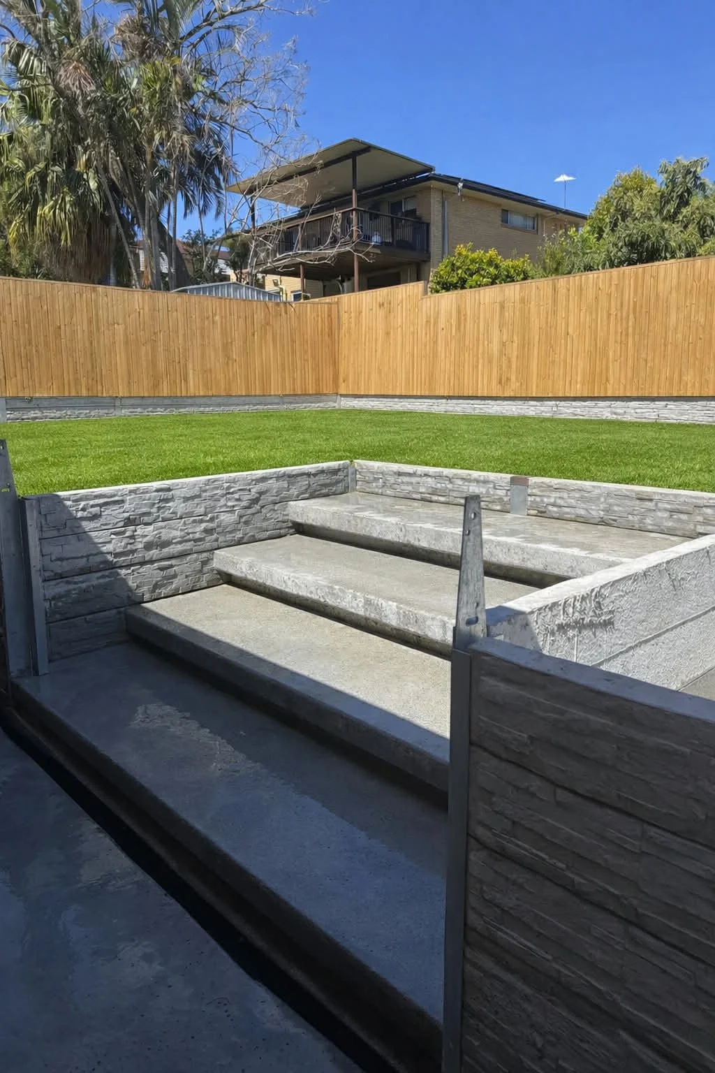 Empty concrete stairs in a backyard with a grassy lawn and a wooden privacy fence, with a house in the background under a clear blue sky.