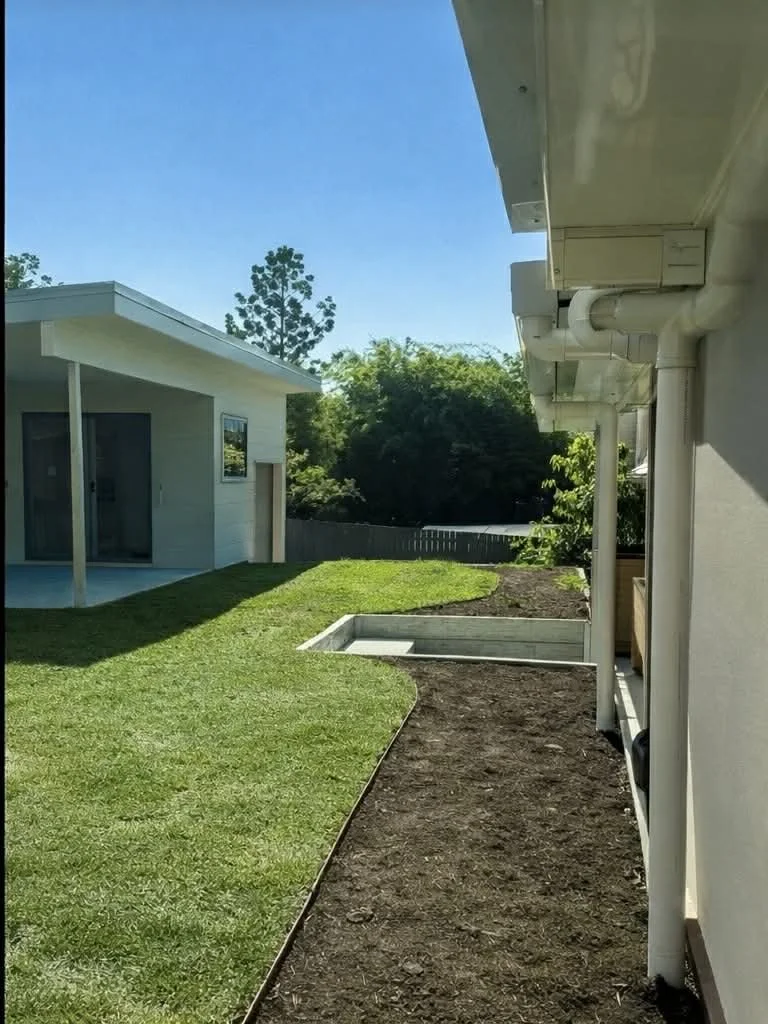 Residential backyard with green grass, part of a house's exterior, and garden bed under construction.