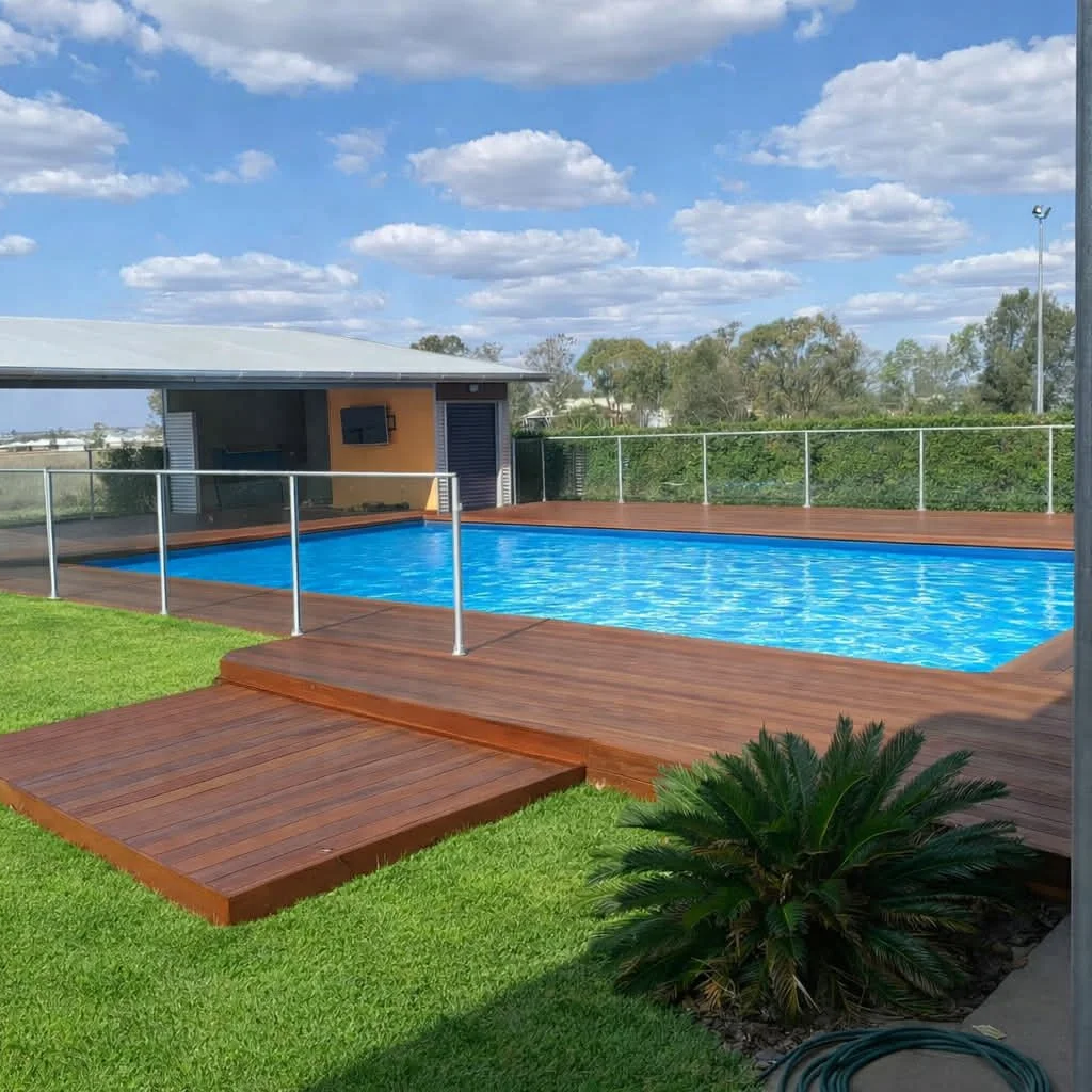 An outdoor swimming pool with a wooden deck, surrounded by a glass fence, under a partly cloudy sky with trees in the background.