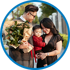 A soldier in uniform holding a small child and talking to a woman outdoors in a park or wooded area.