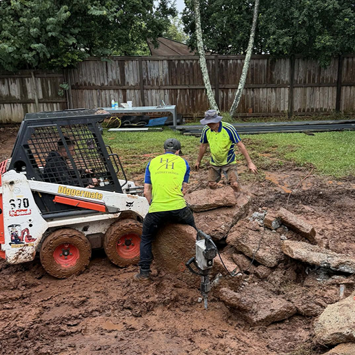 Two construction workers wearing yellow safety vests work on a muddy, uneven ground with large rocks, a small skid steer loader, and a wooden fence in the background.