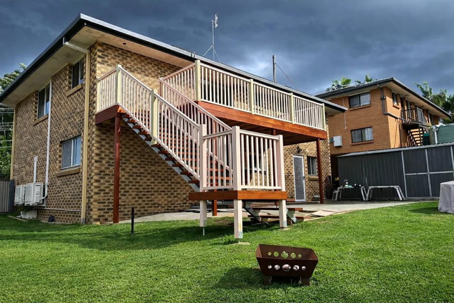 Backyard view of a brick house with an outdoor wooden staircase leading to a second-floor balcony, green lawn, and a dark sky overhead.
