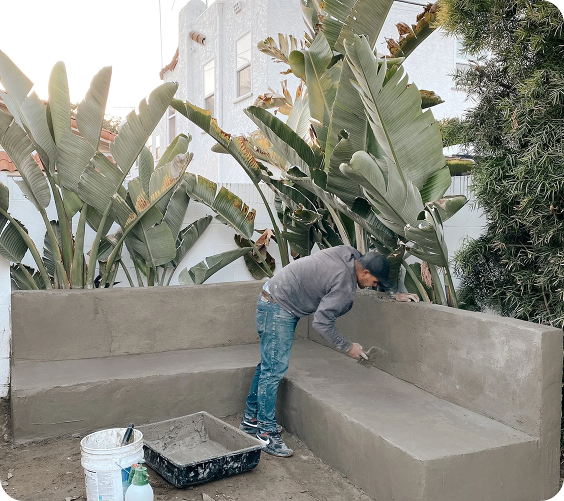 A man building an outdoor built-in concrete bench surrounded by large banana plants and other greenery.
