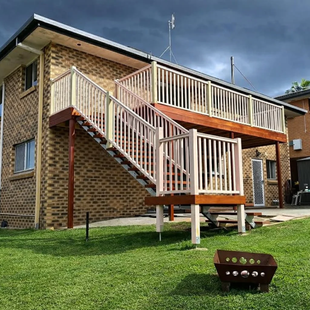 Backyard view of a brick house with a wooden staircase and deck, green lawn, and a rusted metal fire pit, under cloudy sky.