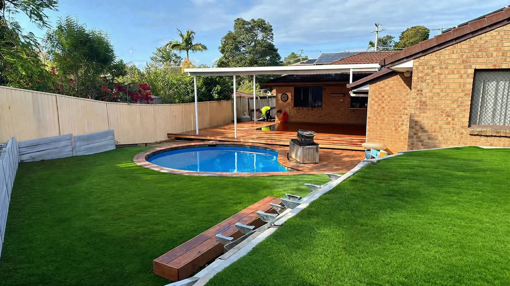 Backyard with a small oval swimming pool, surrounded by green grass and a wooden deck area, with a house and trees in the background.