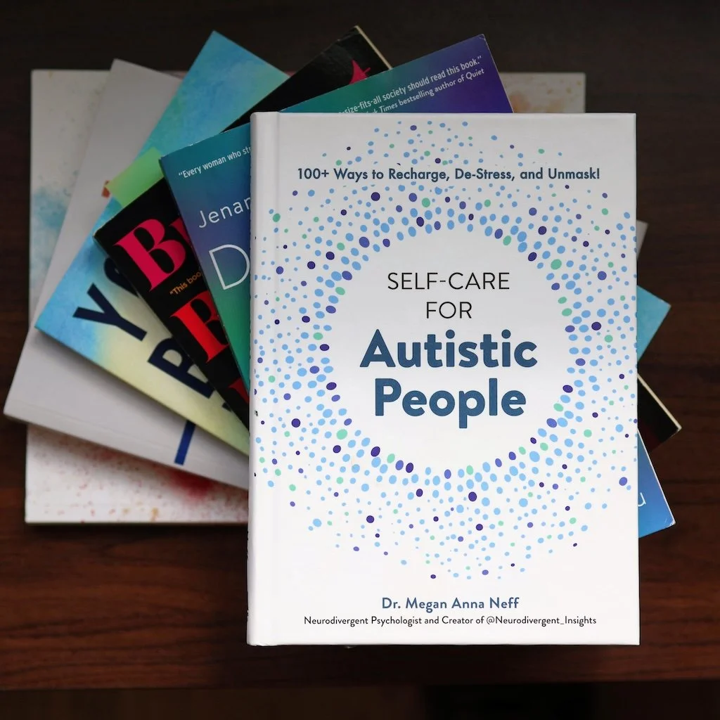 Stack of books with the top book titled 'Self-Care for Autistic People' by Dr. Megan Anna Neff, featuring a white cover with blue and green dot patterns, on a wooden surface.