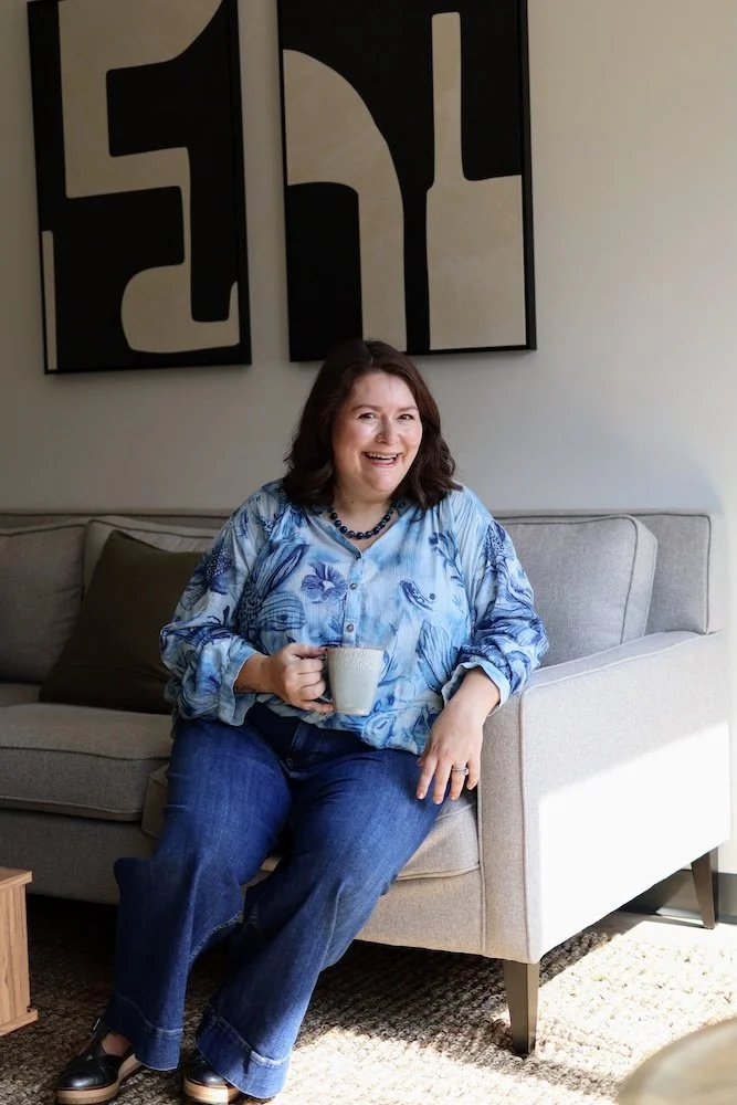 Rebecca Loesberg, LCSW sitting on a light-colored sofa, smiling, holding a white coffee mug, wearing a blue patterned blouse and jeans, with abstract black and white artwork on the wall behind her.