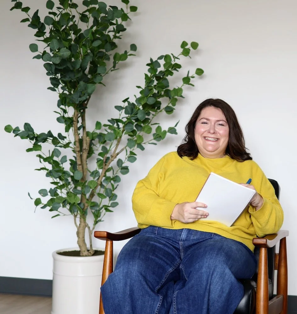 Rebecca Loesberg, LCSW with brown hair wearing a yellow sweater and jeans sitting in a chair beside a large potted green plant, smiling and holding a notepad with a pen.