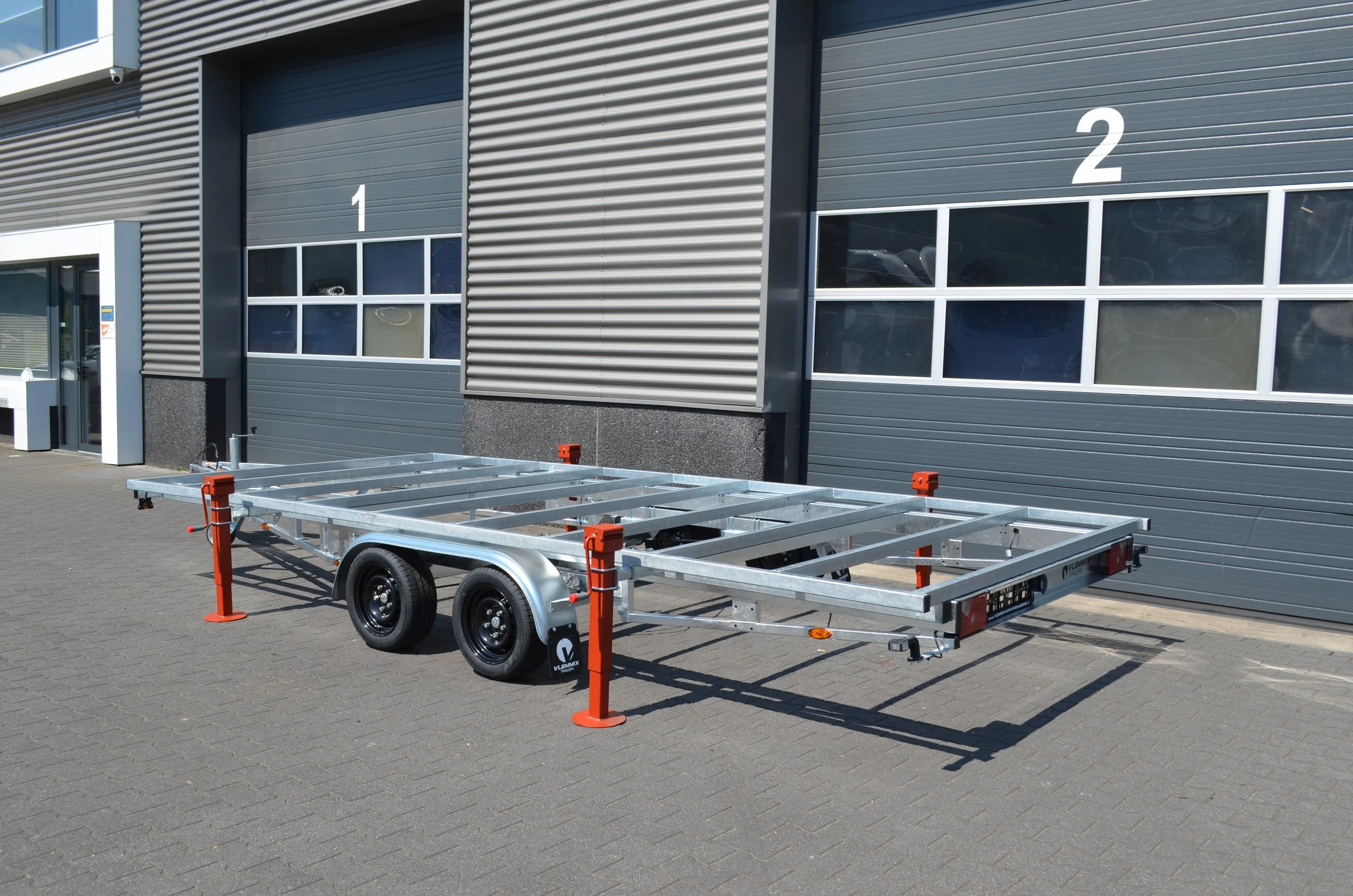A metal utility trailer with two axles parked on a paved area in front of a building with grey shutter doors. One door is labeled '1' and the other '2'. The trailer has no sides or cargo, and orange safety barriers are placed around it.
