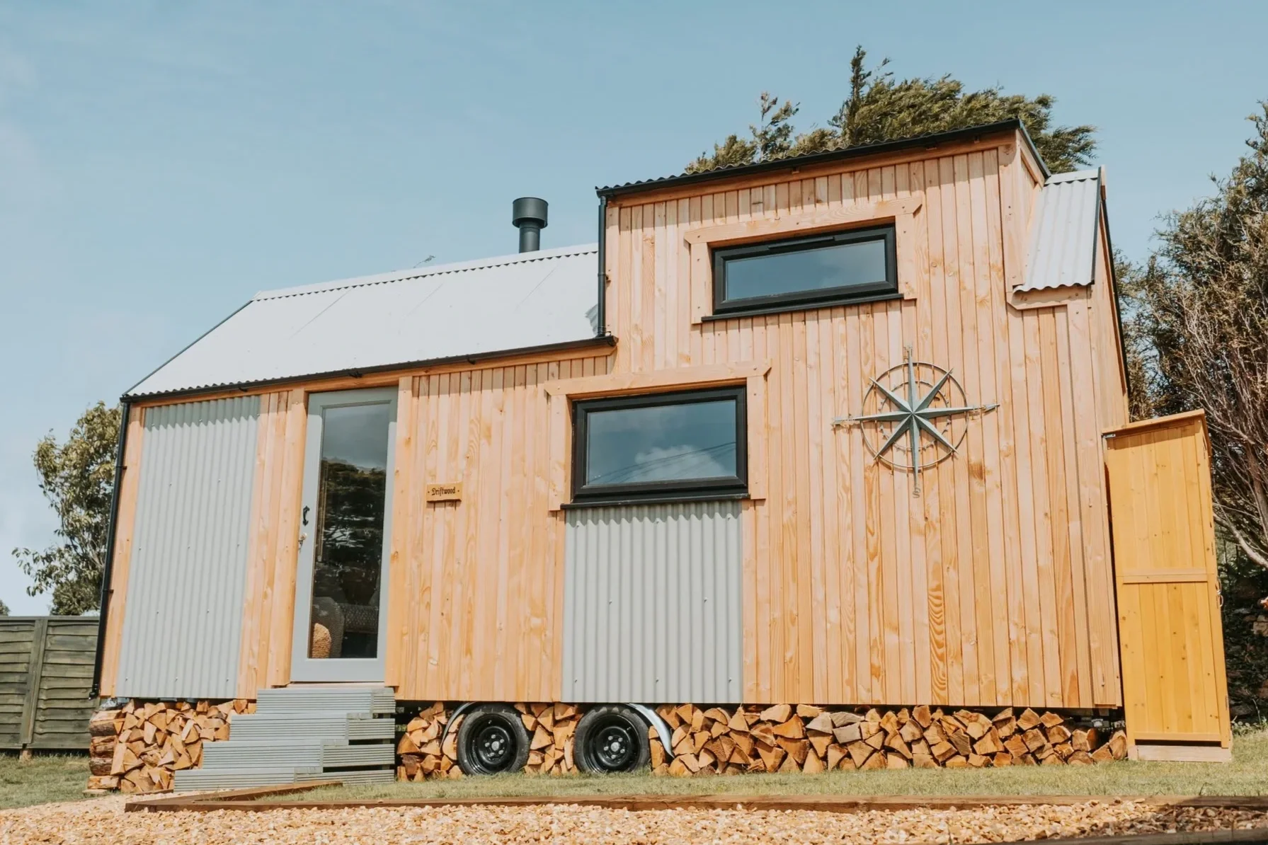 A tiny house with wooden and corrugated metal siding, set on a bed of cut firewood, featuring black-framed windows, a small staircase, and a metal sunburst decoration on the front.