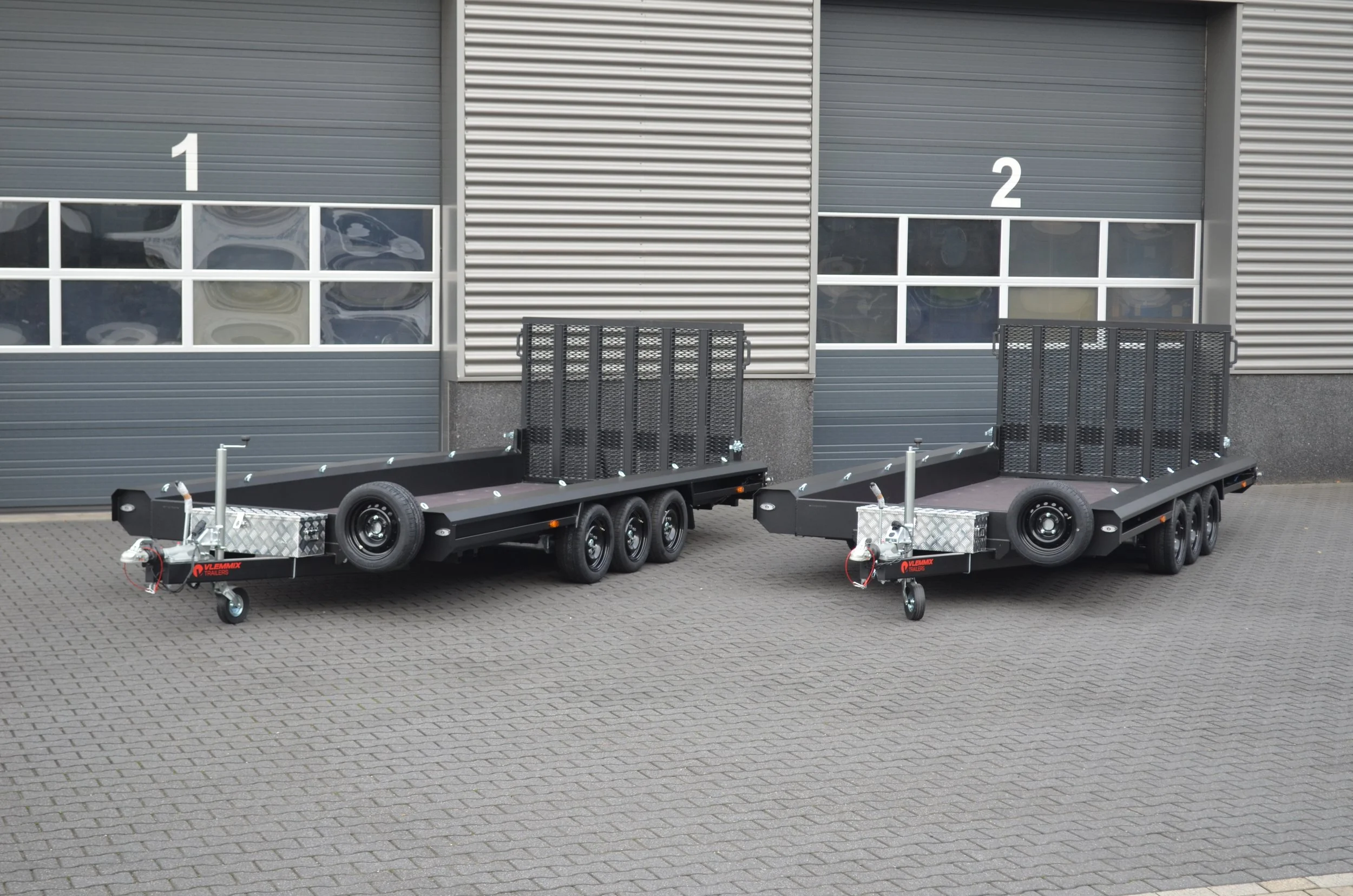 Two black flatbed trailers parked side by side in front of a corrugated metal building with large windows labeled '1' and '2'.