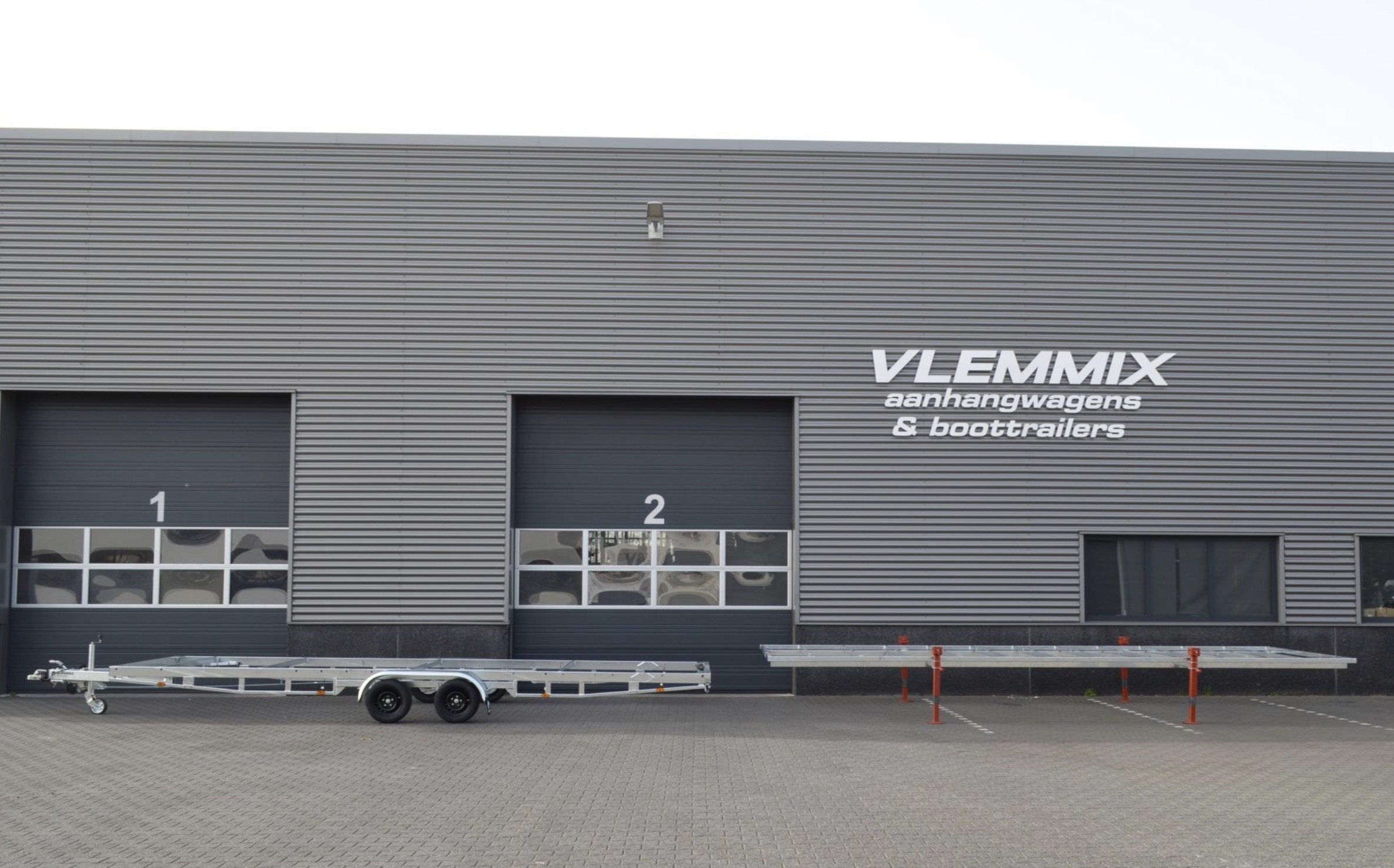 A modern industrial building with metal siding, featuring two large garage doors numbered 1 and 2, and a sign that reads 'Vlemmix aanhangwagens & boottrailers.' A flatbed trailer is parked in front of the building.