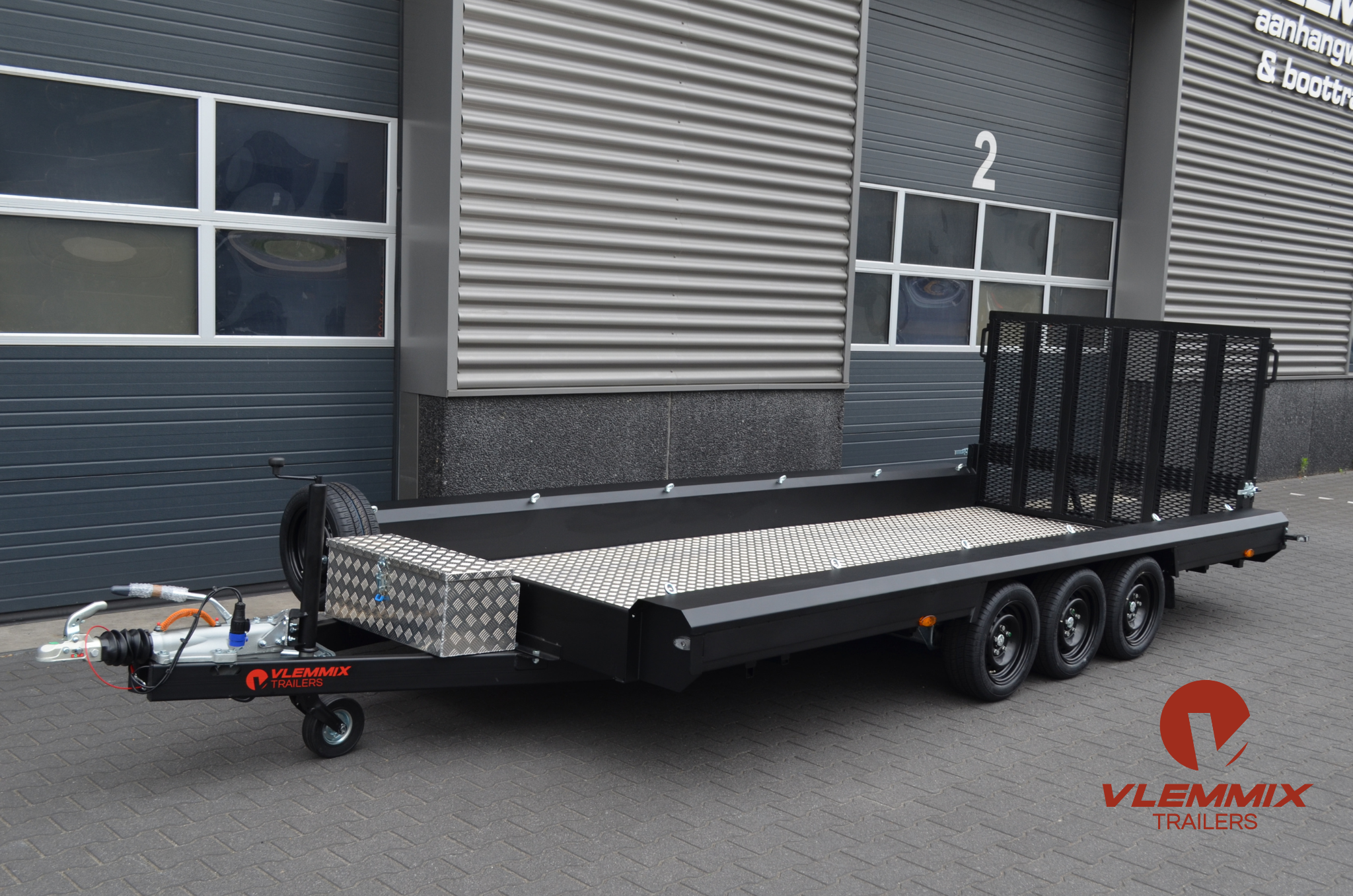 Black flatbed trailer with a diamond plate surface, labeled 'Vlemmix Trailers' in red, parked on a paved surface in front of a modern industrial building with gray metal siding and windows.