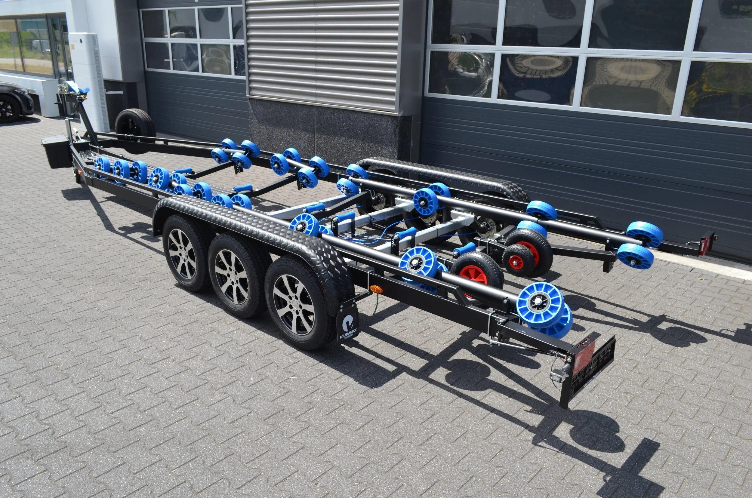 A black vehicle trailer with multiple blue rollers, parked outside a gray industrial building with large windows.