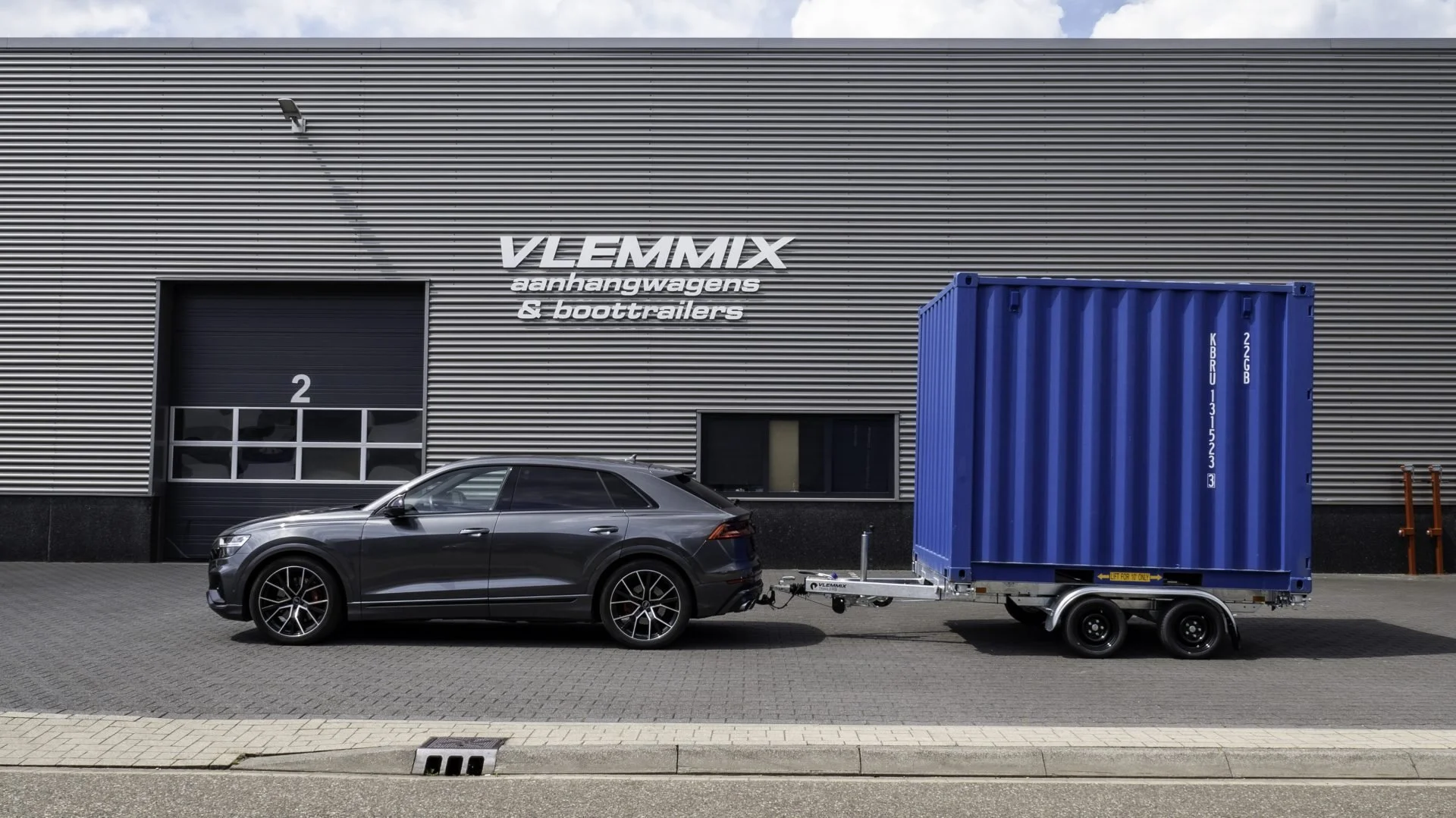 A gray car attached to a trailer with a blue container parked outside a building labeled Vlemmix aanhangwagens & boottrailers.