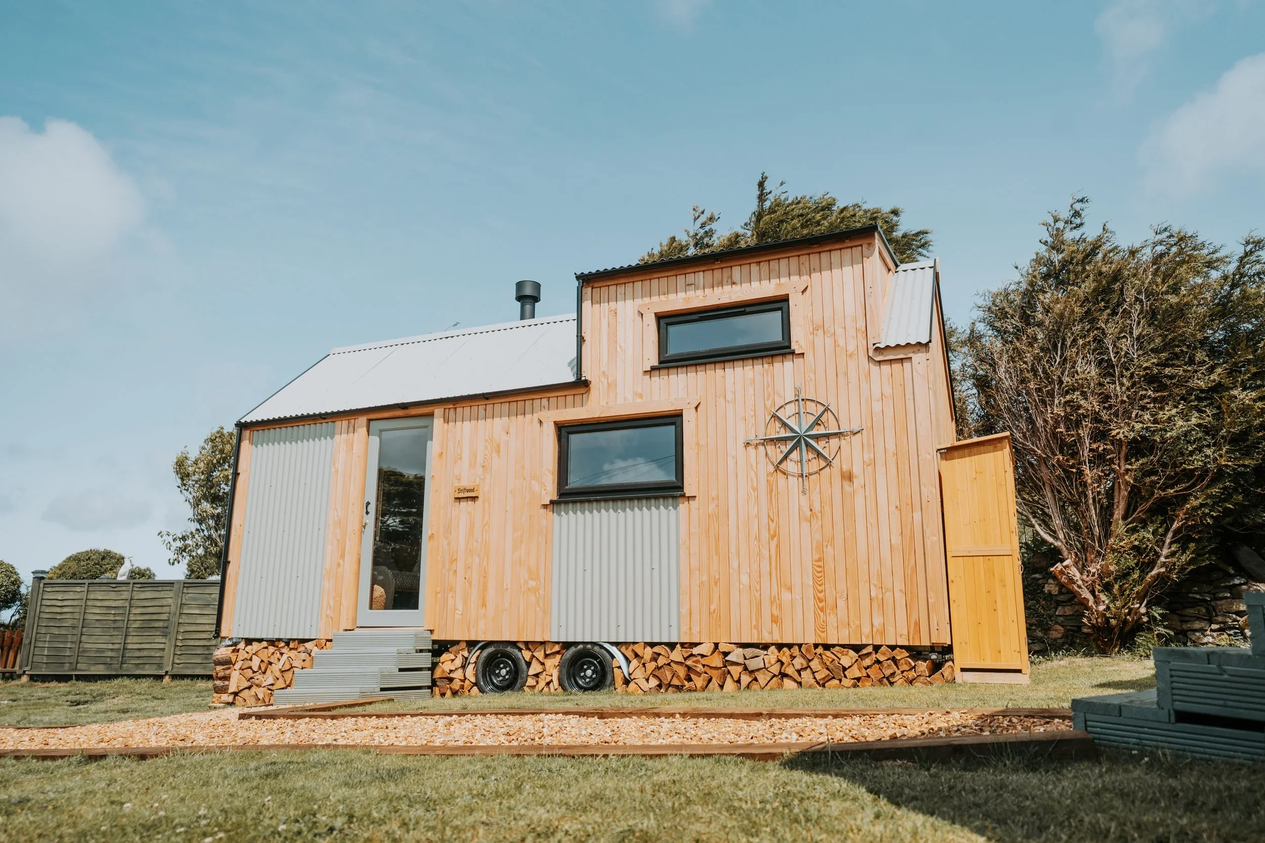A modern tiny house built on wheels with wooden and metal exterior, featuring large windows, a metal roof, and a decorative wheel on the side. It is parked on a grassy area with trees in the background.