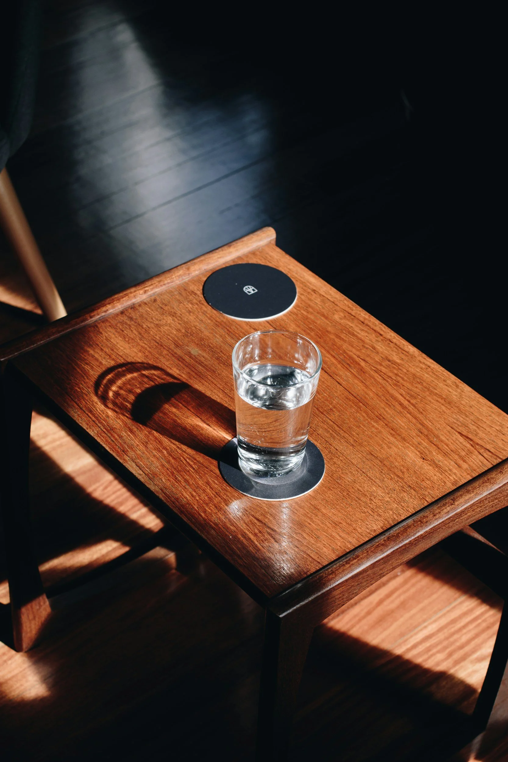 A glass of water on a wooden table with a black circular wireless charging pad nearby, casting a shadow on the surface.