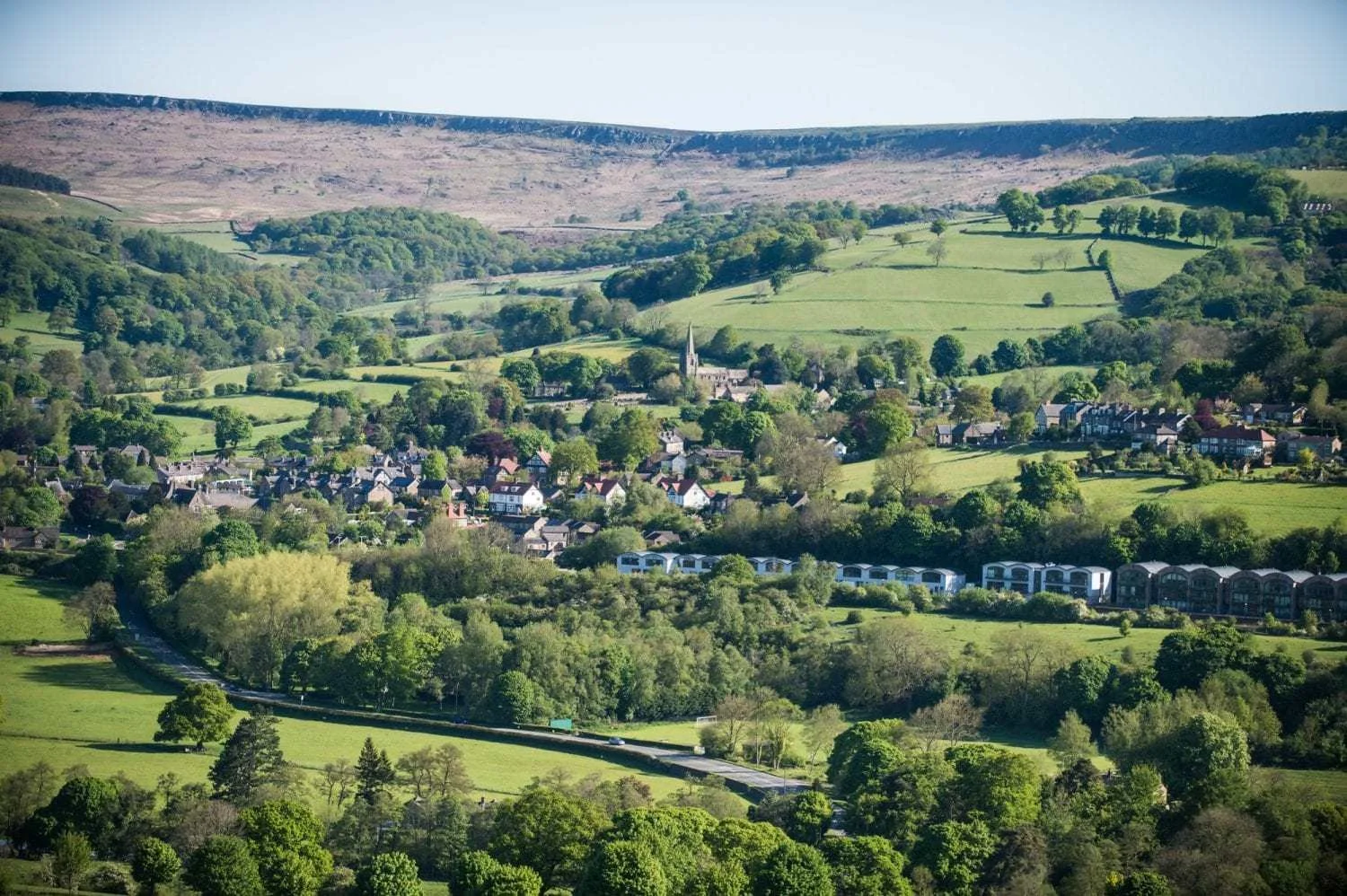 Scenic view of a small town surrounded by lush green hills and trees, with a winding road and various residential buildings, under a clear blue sky.