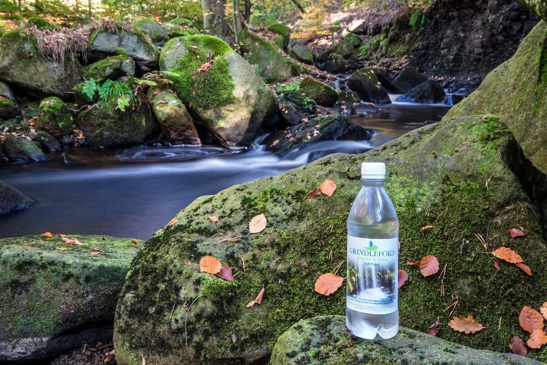 A clear plastic water bottle with a label reading "Grindleford" in front of a mossy rock in a forest stream, with autumn leaves scattered on the rocks.