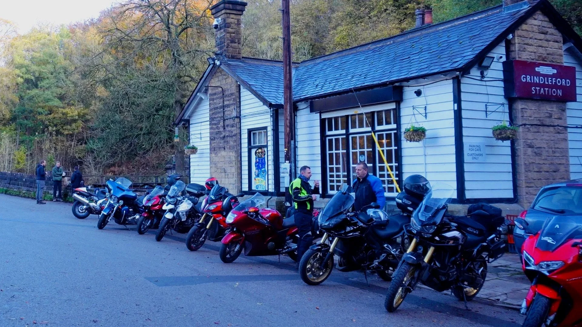 A small train station named Grindleford Station with a group of motorcycles parked in front. Two men are standing and talking near the motorcycles. There are three women standing to the left side of the station building. The station is located near a wooded area with fall foliage.