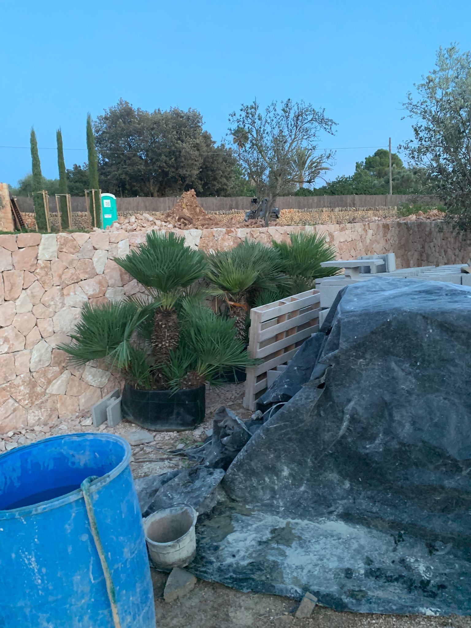 Construction site with a stone wall, trees, construction materials, piles of dirt, and buckets in the foreground during dusk.