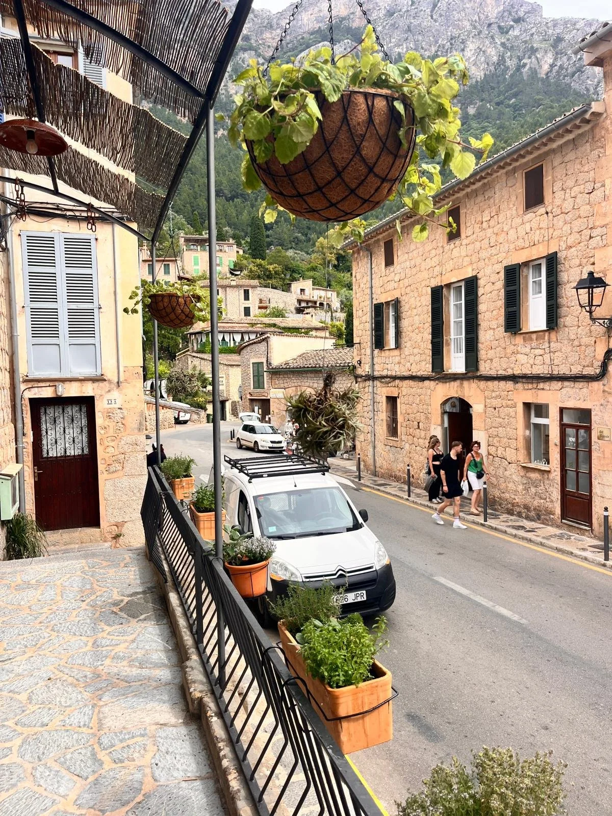 Street scene in a European village with stone buildings, parked cars, and pedestrians walking along the sidewalk. Hanging flower baskets and potted plants decorate the area.