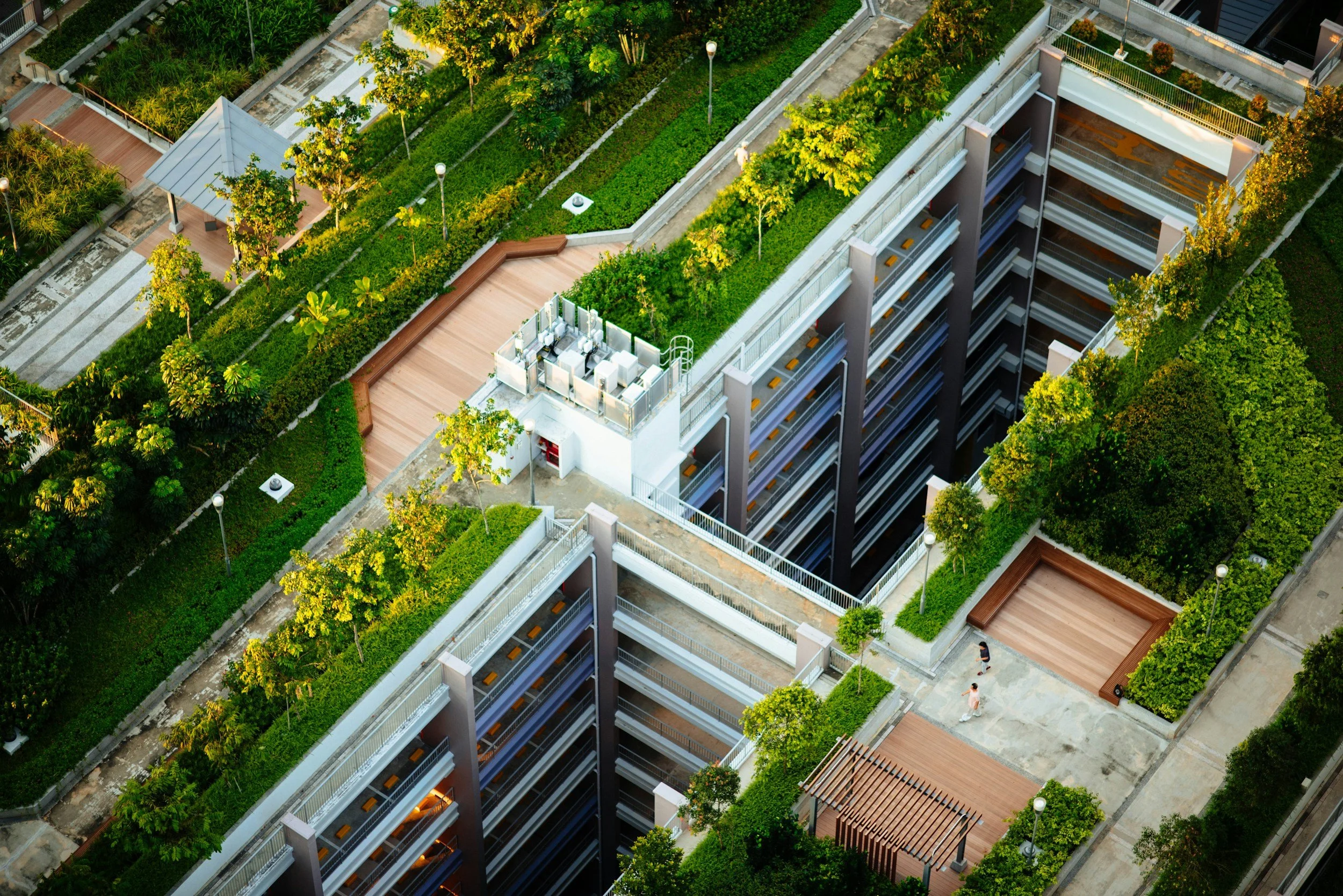 Aerial view of a multi-story building with rooftop gardens, walkways, and trees, with people walking on pathways.