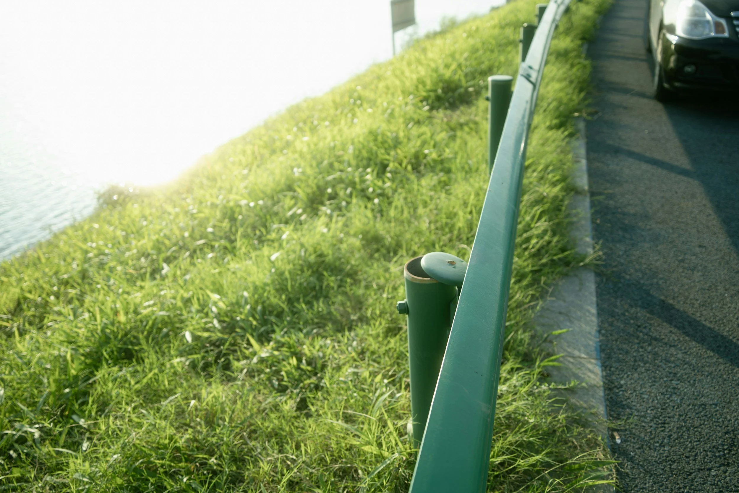 Green metal railing beside a paved road with green grass and a car parked to the side.