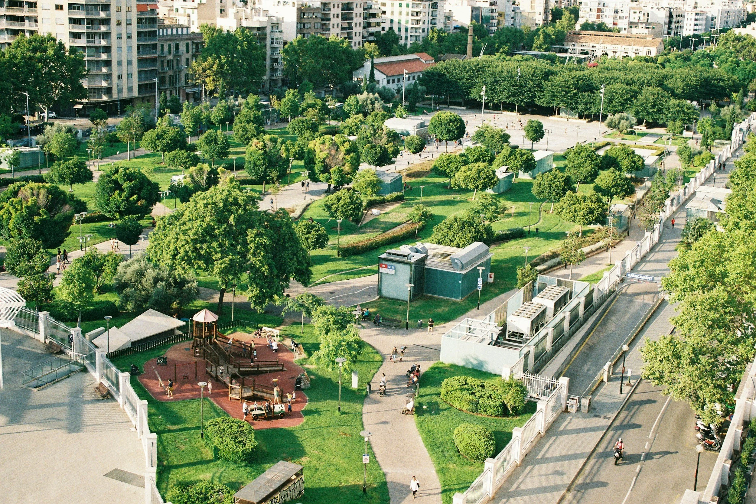 An aerial view of a city park with green trees, walking paths, and people enjoying leisure activities. Surrounding the park are tall residential buildings, and in the foreground, there's a playground area with children and adults.